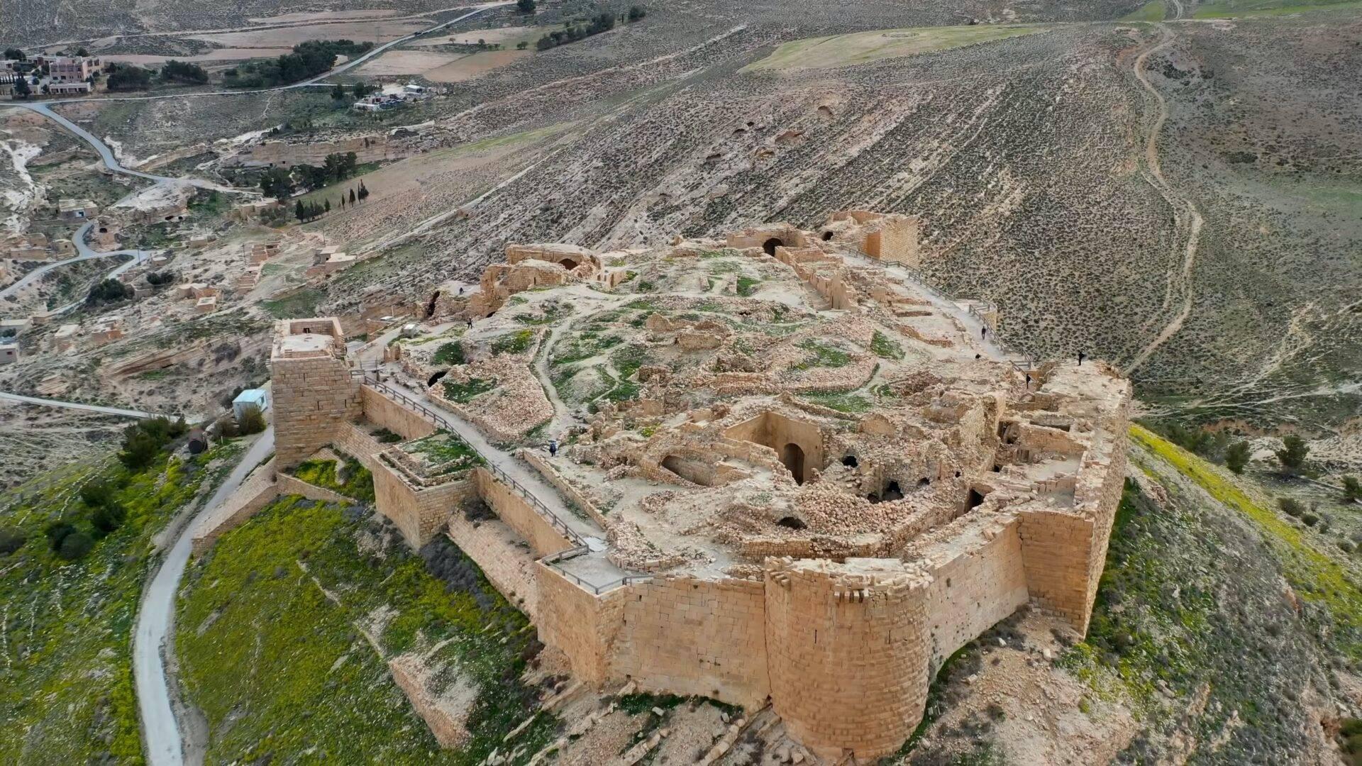Forteresse en ruines vue d’en haut avec murs de pierre, dominating un paysage montagneux aride et vallée verdoyante en arrière-plan