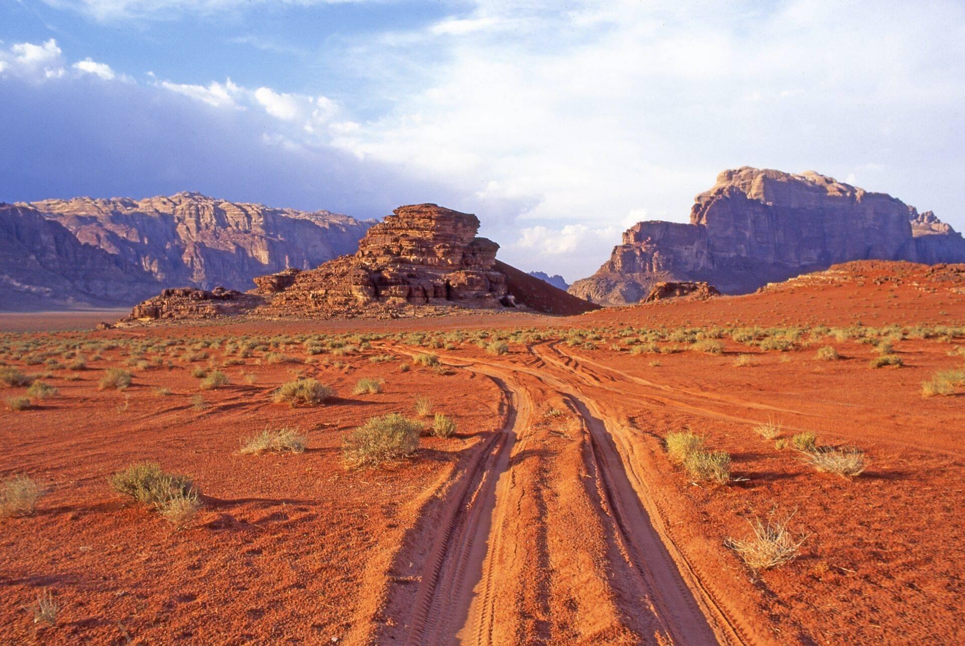 Route de terre rouge traversant un désert aride avec formations rocheuses monumentales sous un ciel bleu nuageux