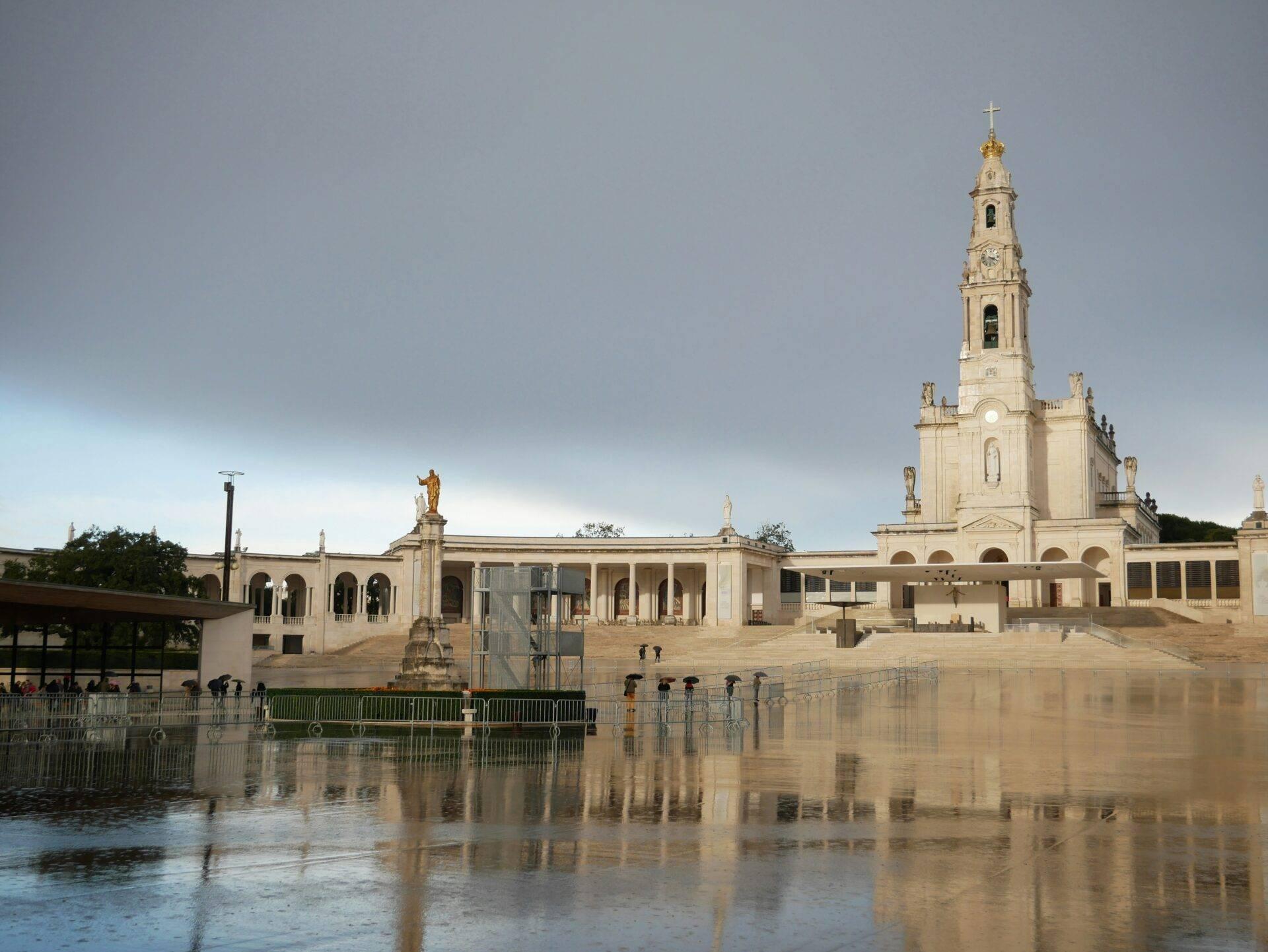 Un sanctuaire religieux blanc avec campanile dominant une esplanade pavée bordée d’eau calme et réfléchissante