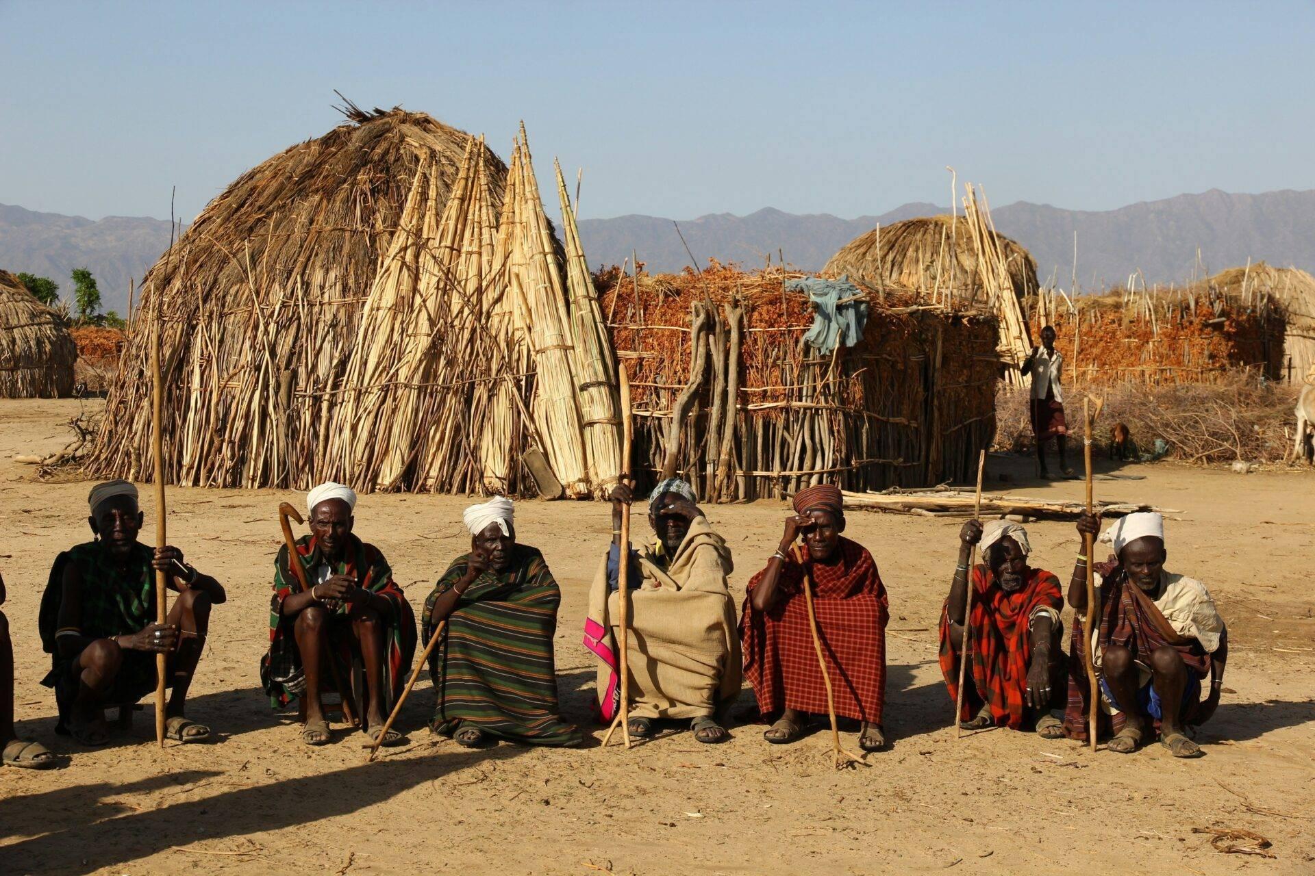 Groupe de guerriers éthiopiens en tenues traditionnelles colorées posant devant des huttes en paille dans un village pastoral