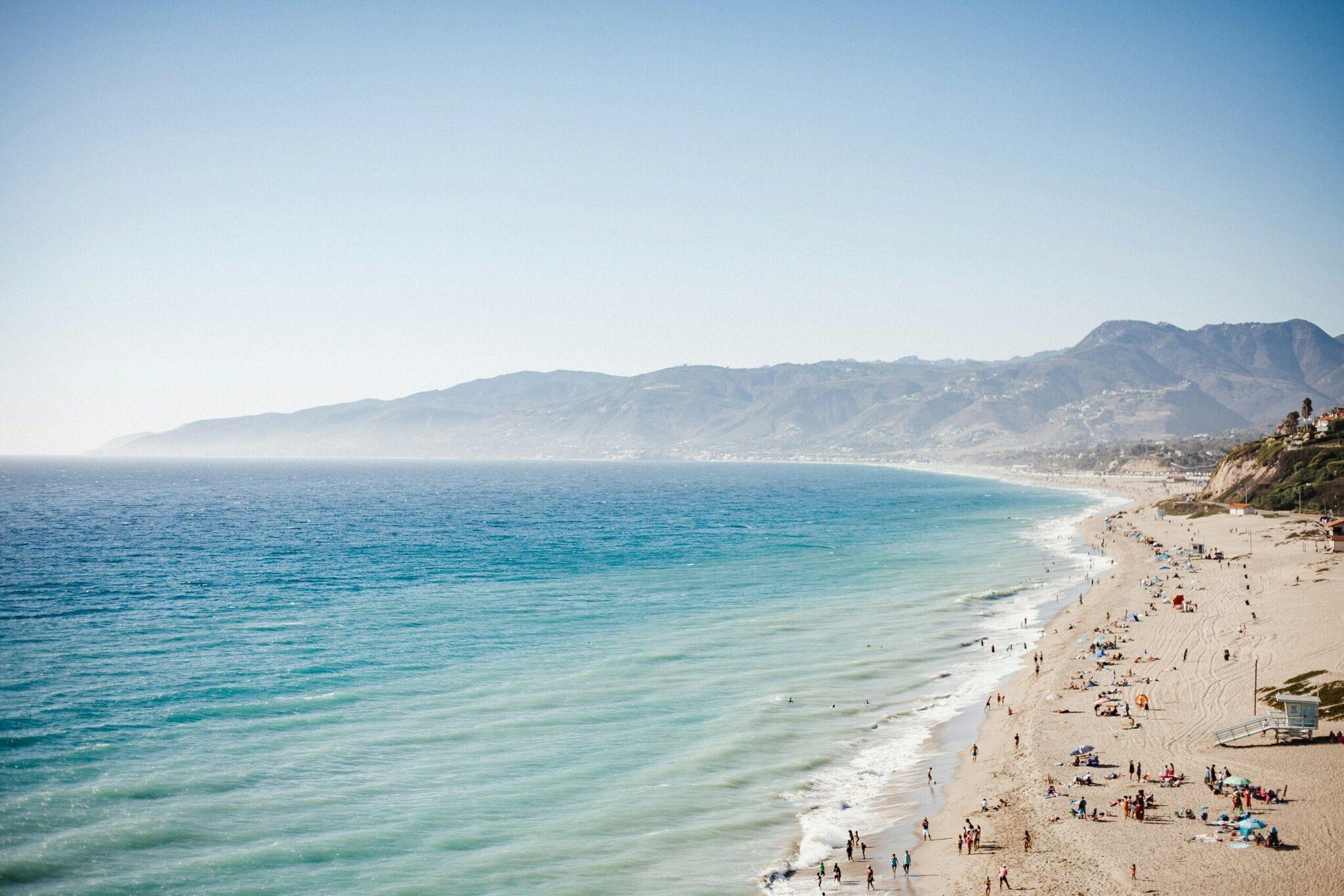 Une longue plage de sable blanc avec baigneurs, face à un océan turquoise et des montagnes côtières boisées