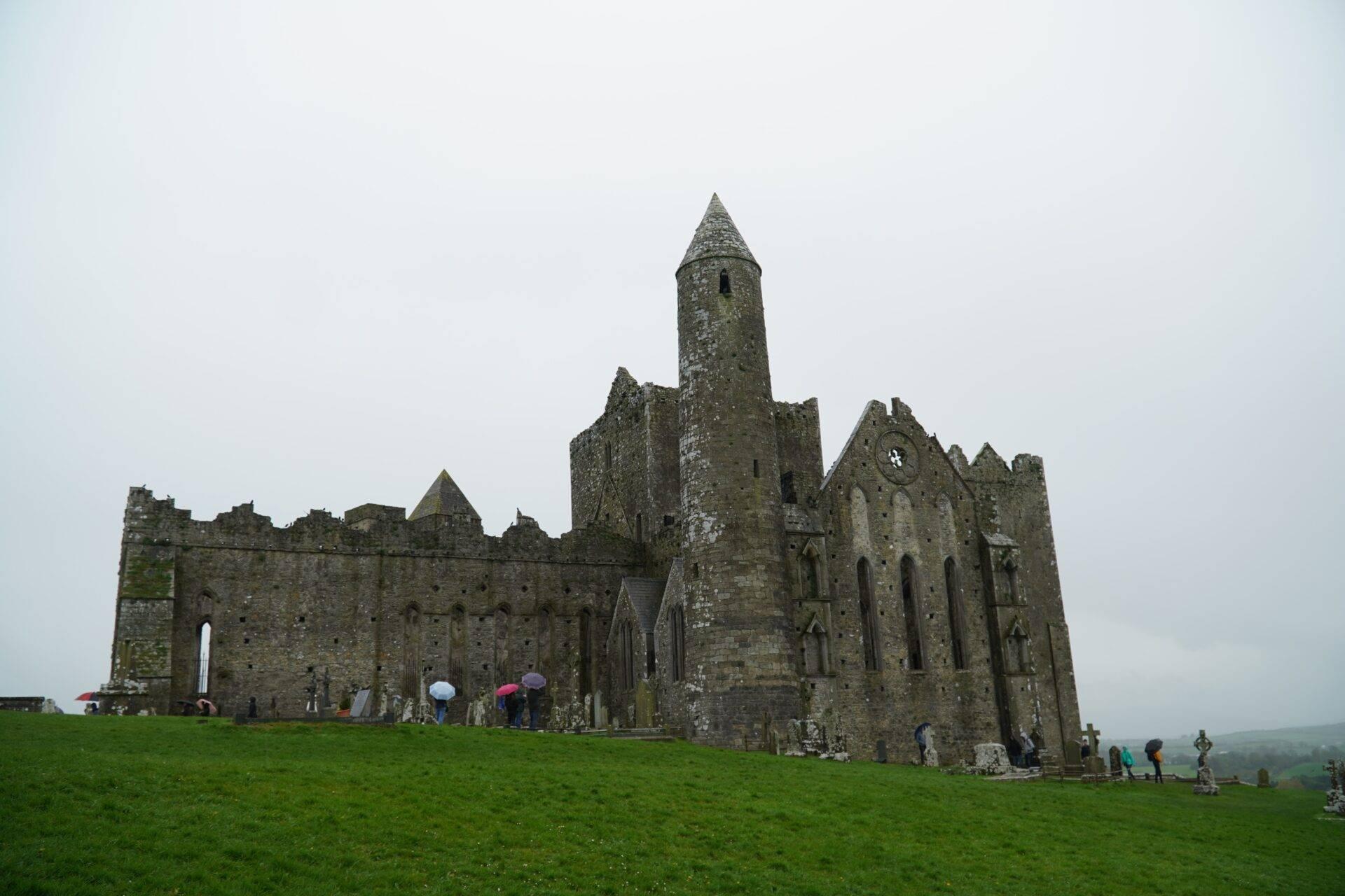 Ruines d’une abbaye médiévale en pierre avec tour ronde et cimetière, sous ciel nuageux gris