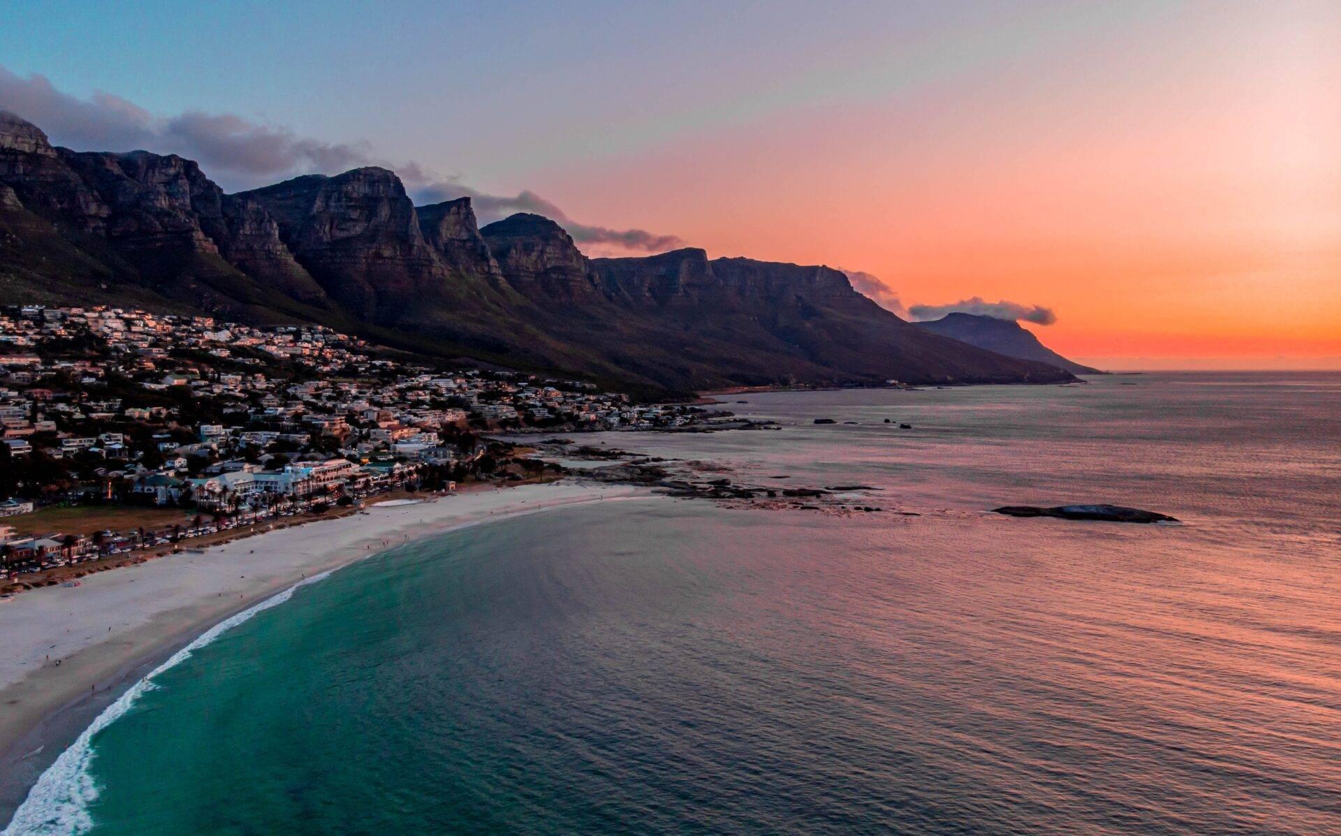 Coucher de soleil sur une plage de sable blanc face à des montagnes rocheuses majestueuses surplombant une baie côtière