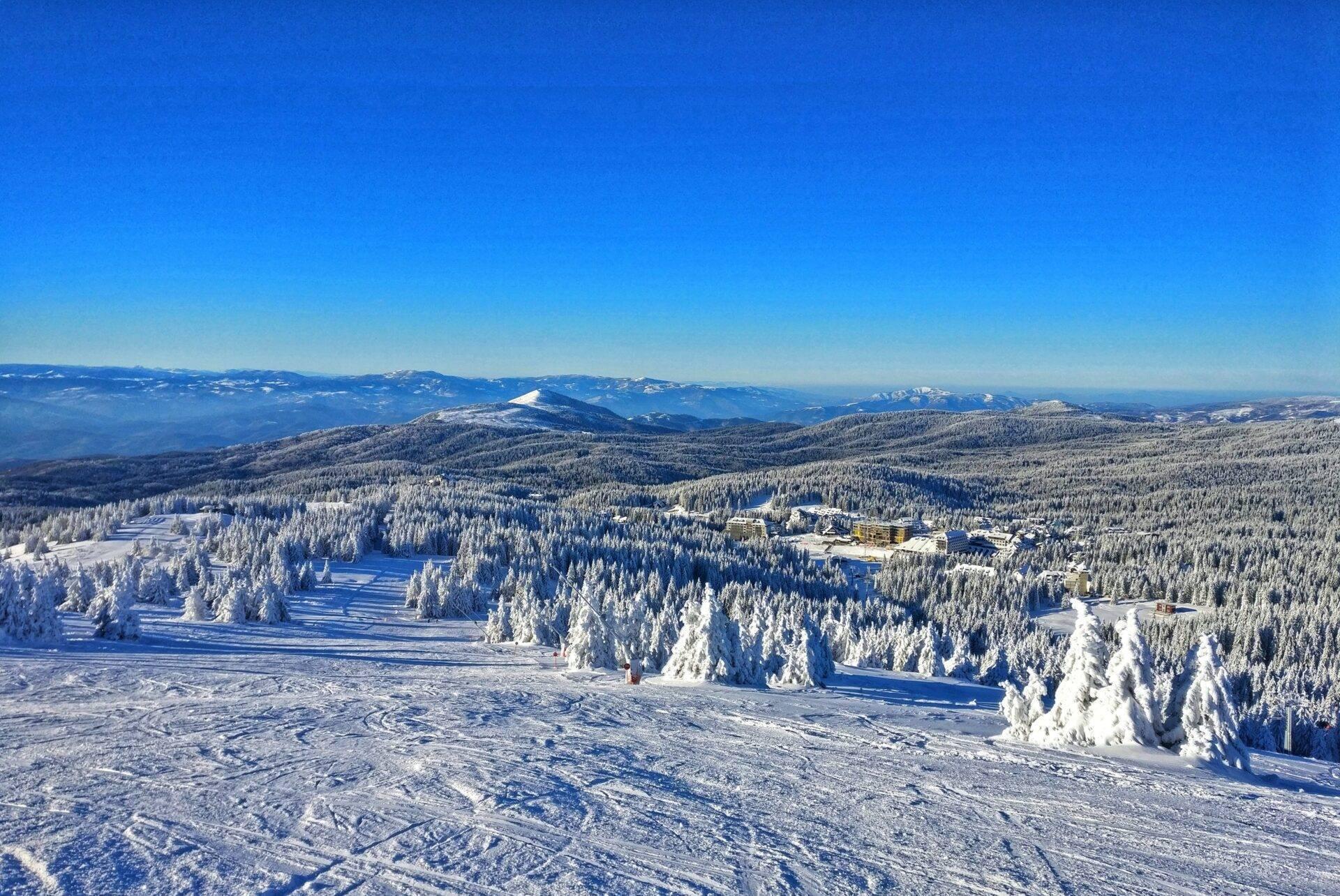 Vue aérienne d’une région montagneuse enneigée avec forêts de conifères, lacs gelés et bâtiments en fond de vallée