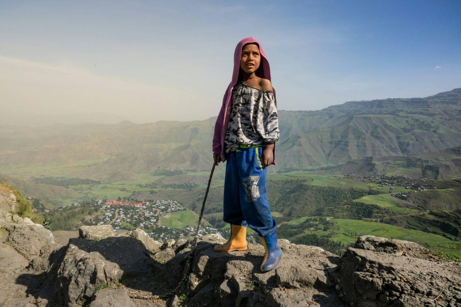 Une jeune fille en tenue traditionnelle pose sur les hauteurs rocheuses d’une vallée verdoyante et montagneuse