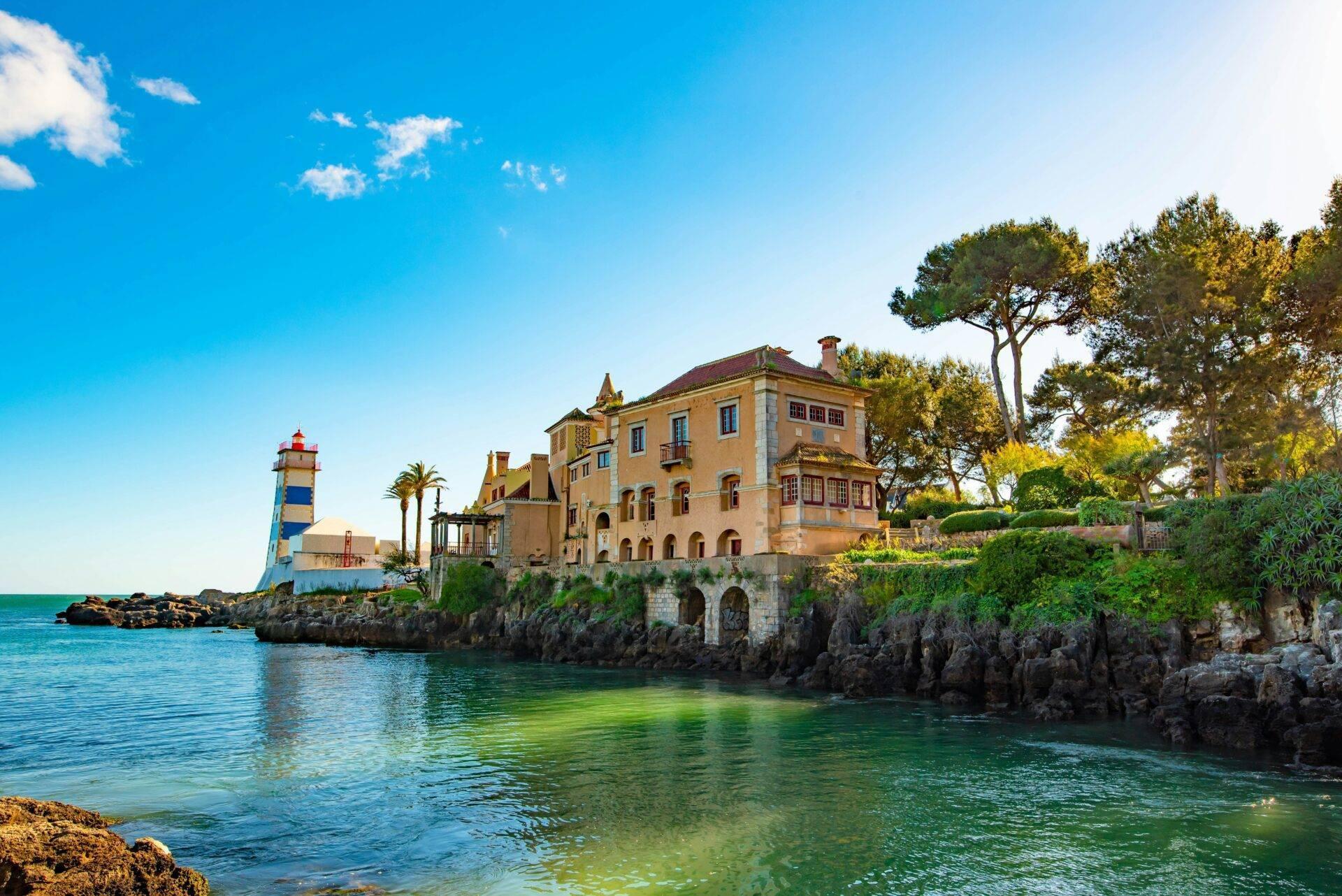 Un palais ocre perché sur des rochers côtiers avec un phare bleu et blanc, surplombant une baie turquoise méditerranéenne