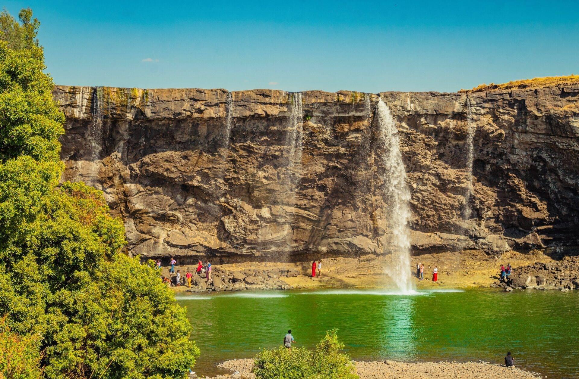 Une cascade spectaculaire jaillit d’une falaise rocheuse dans un lac turquoise, entourée de visiteurs et de végétation verdoyante