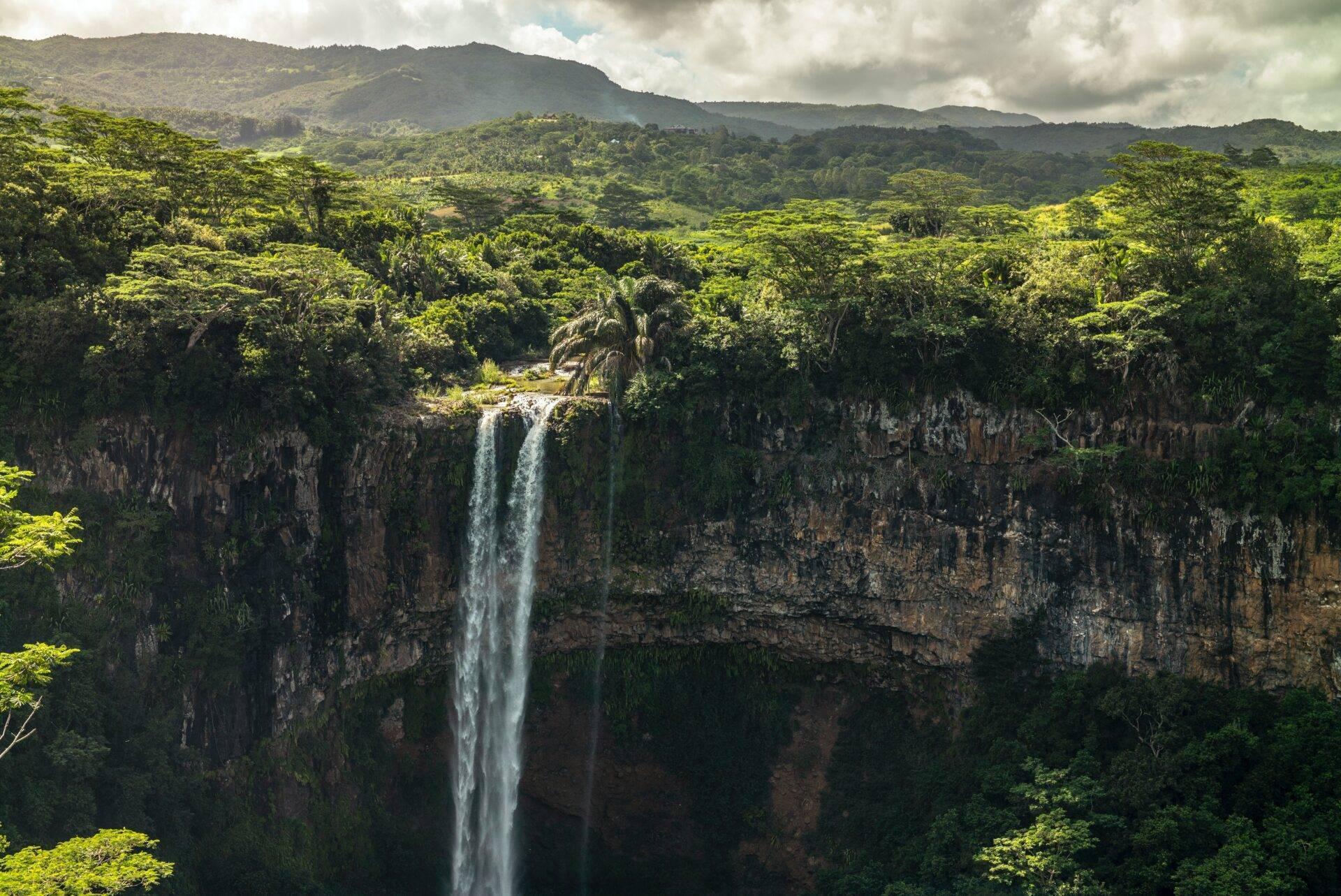 Une cascade spectaculaire s’écoule d’une falaise basaltique dans une vallée tropicale luxuriante entourée de montagnes boisées