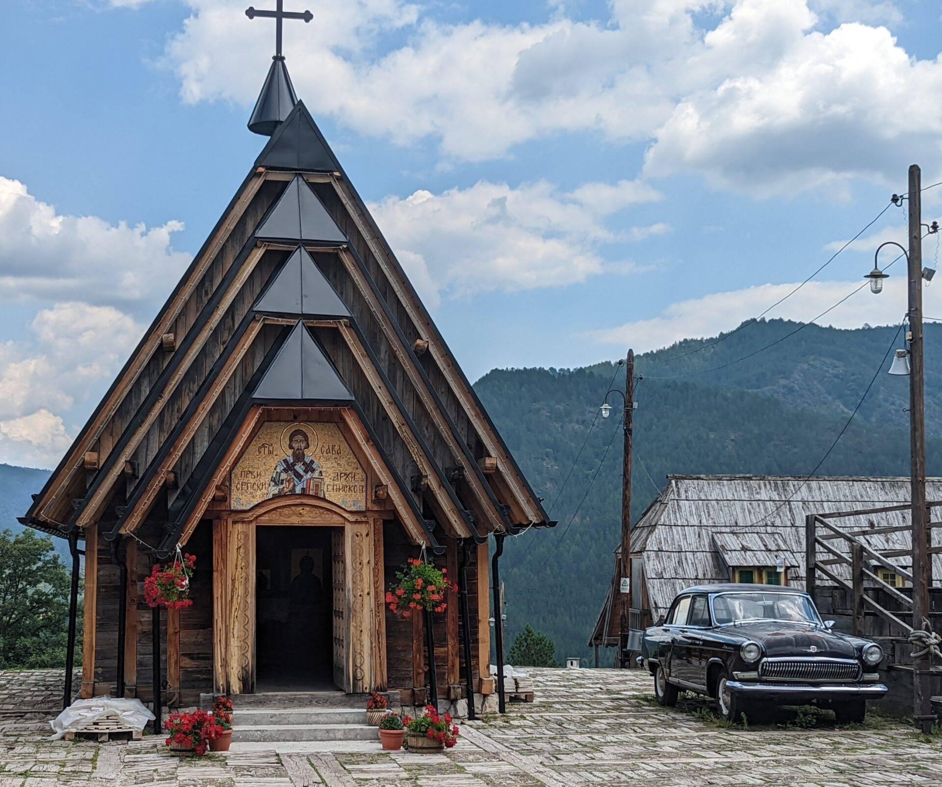 Une petite église orthodoxe en bois avec croix dorée, voiture vintage et montagnes boisées en arrière-plan