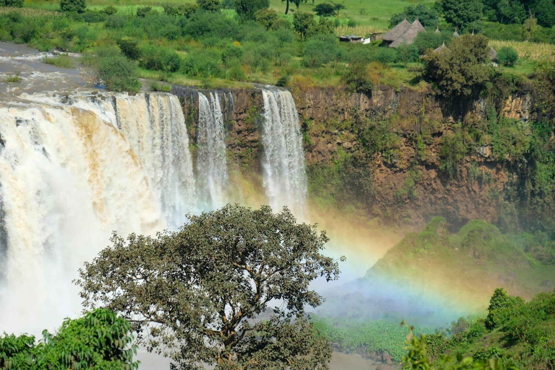 Spectaculaires chutes d’eau en cascade sur falaise rocheuse avec arc-en-ciel, végétation luxuriante et habitations traditionnelles en arrière-plan