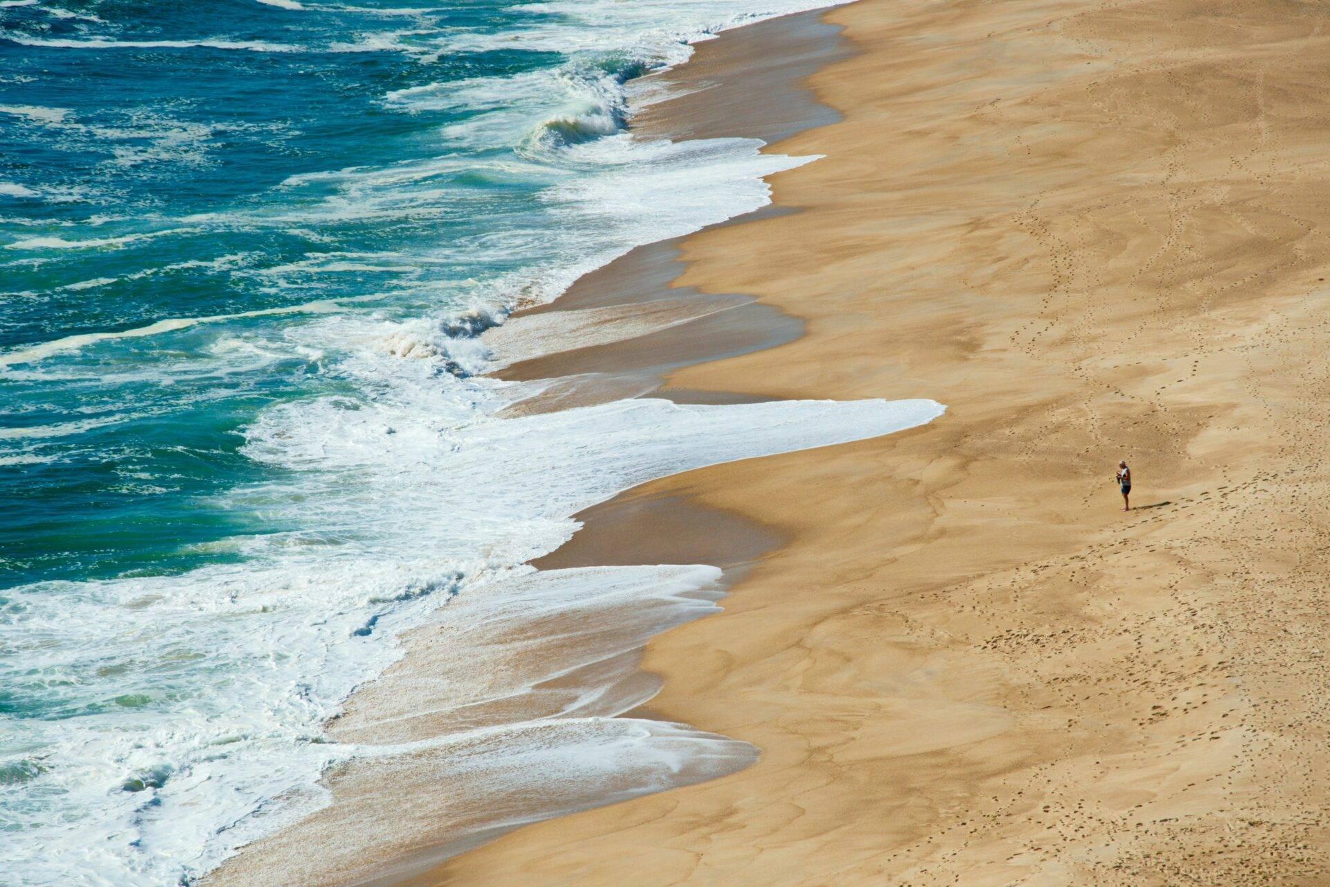 Vue aérienne d’une plage de sable doré avec vagues déferlantes et un marcheur solitaire au Portugal