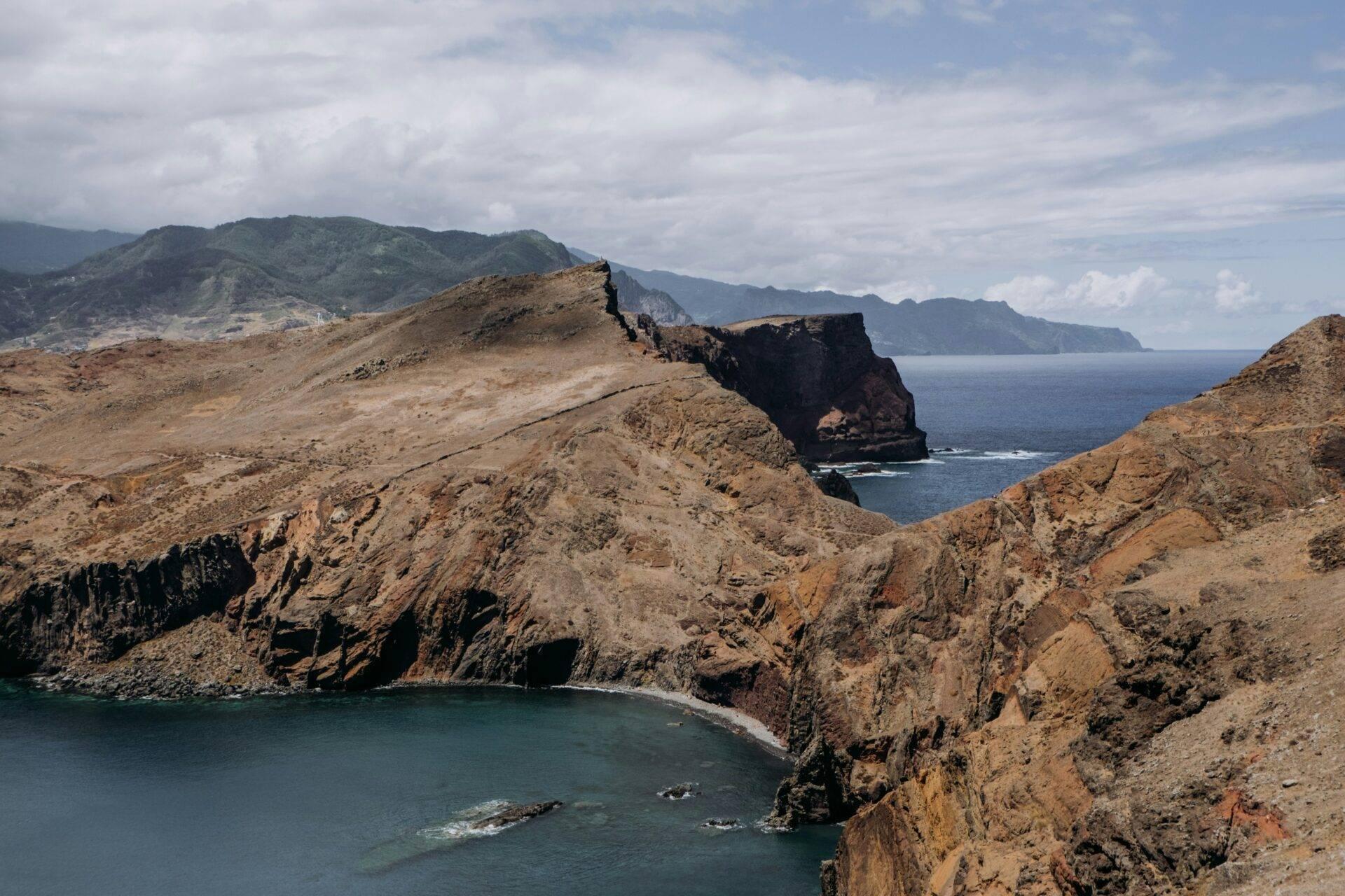 Paysage côtier aride avec falaises rocheuses, petite baie turquoise et îles volcaniques en arrière-plan sous ciel nuageux