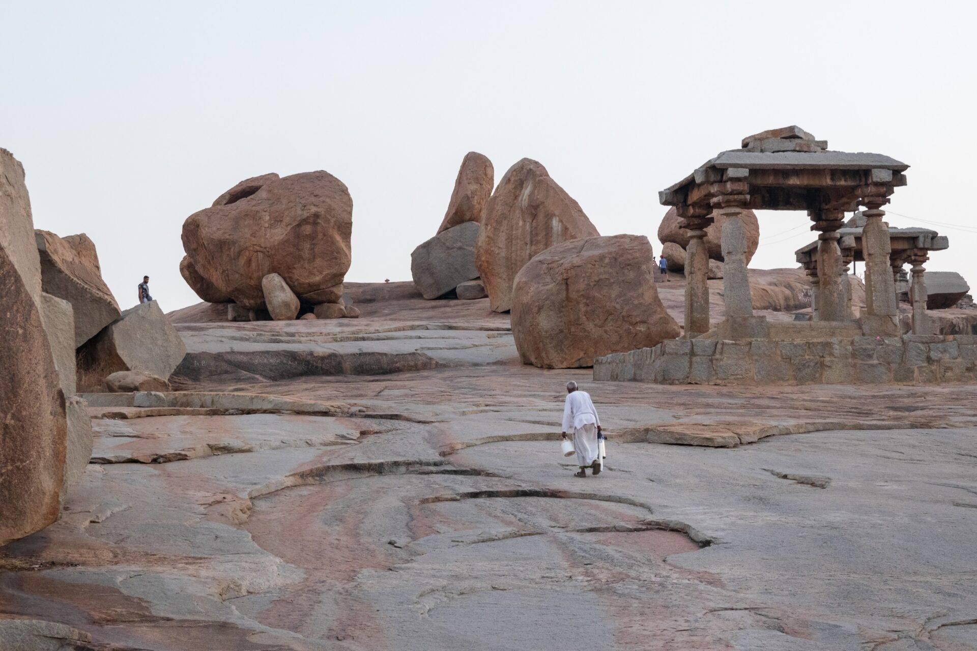 Un homme en vêtements blancs marche sur un plateau rocheux parsemé de boulders géants et de ruines de temples en pierre