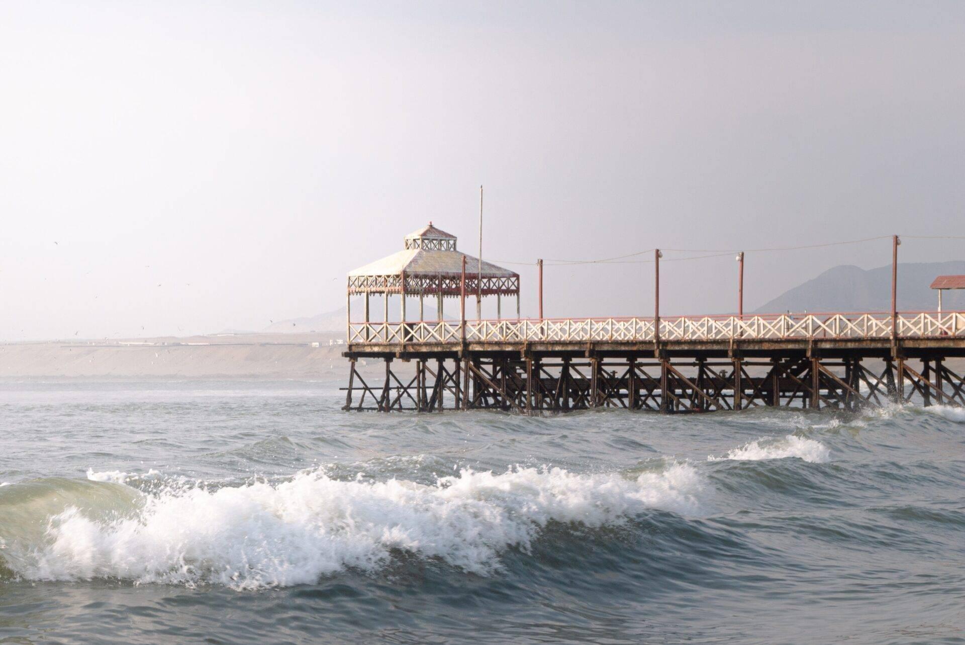 Un pier en bois avec un pavillon octogonal face à l’océan avec des vagues et des montagnes en arrière-plan