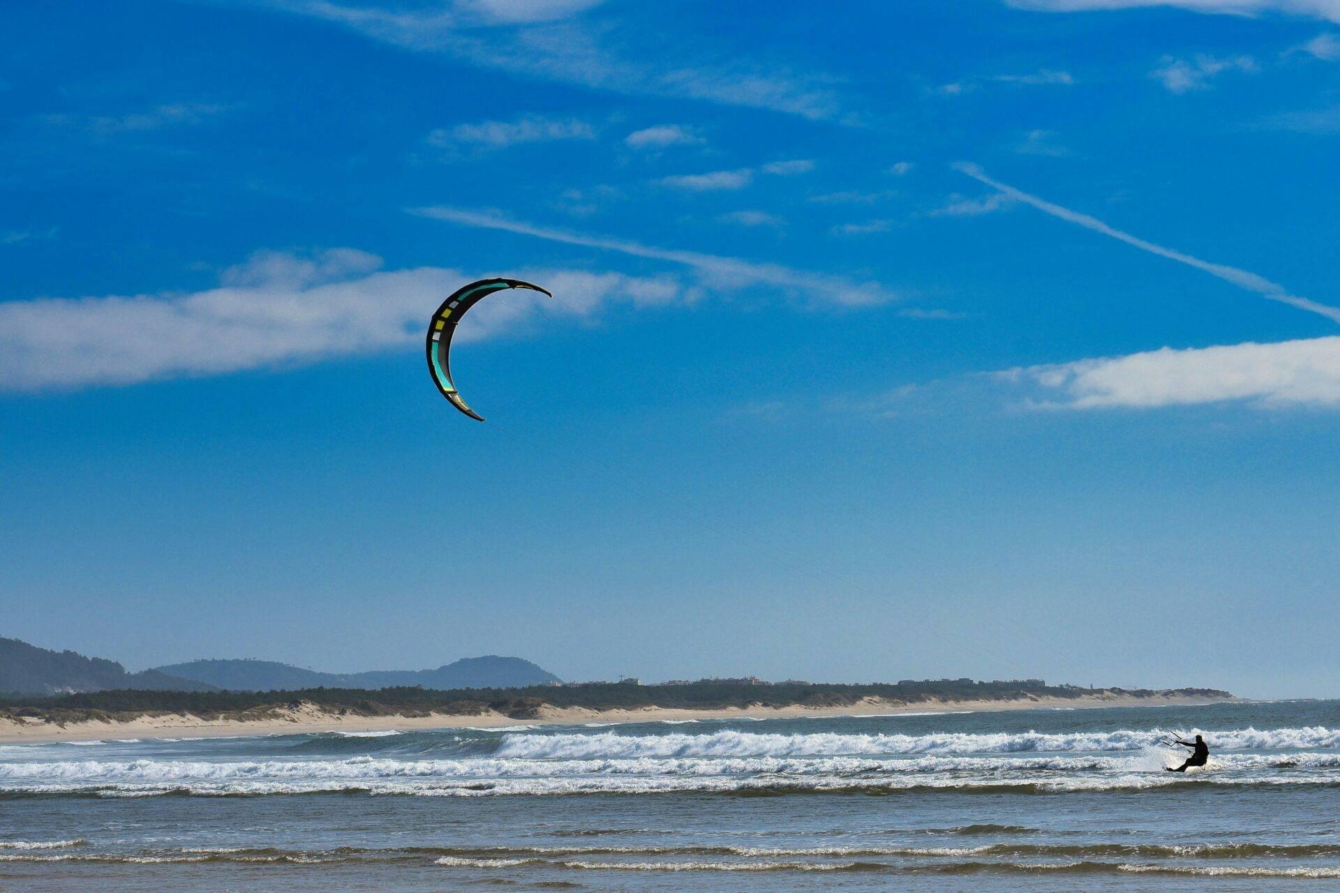 Un kitesurfeur en action sur une plage de sable fin avec des vagues, sous un ciel bleu dégagé et des montagnes en arrière-plan