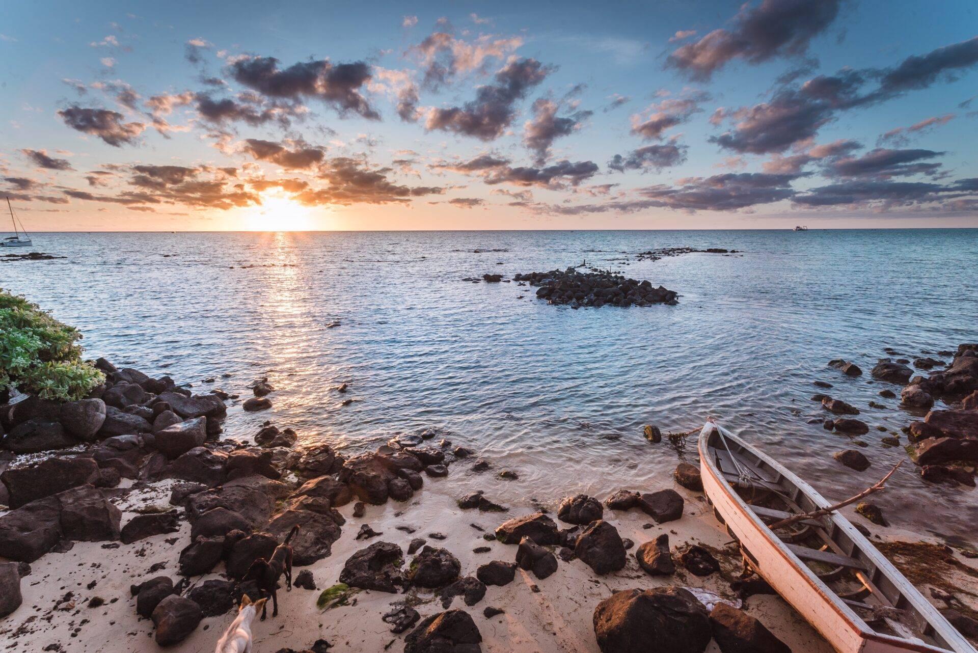 Coucher de soleil sur une plage rocheuse avec deux barques de pêche échouées et voiliers en arrière-plan