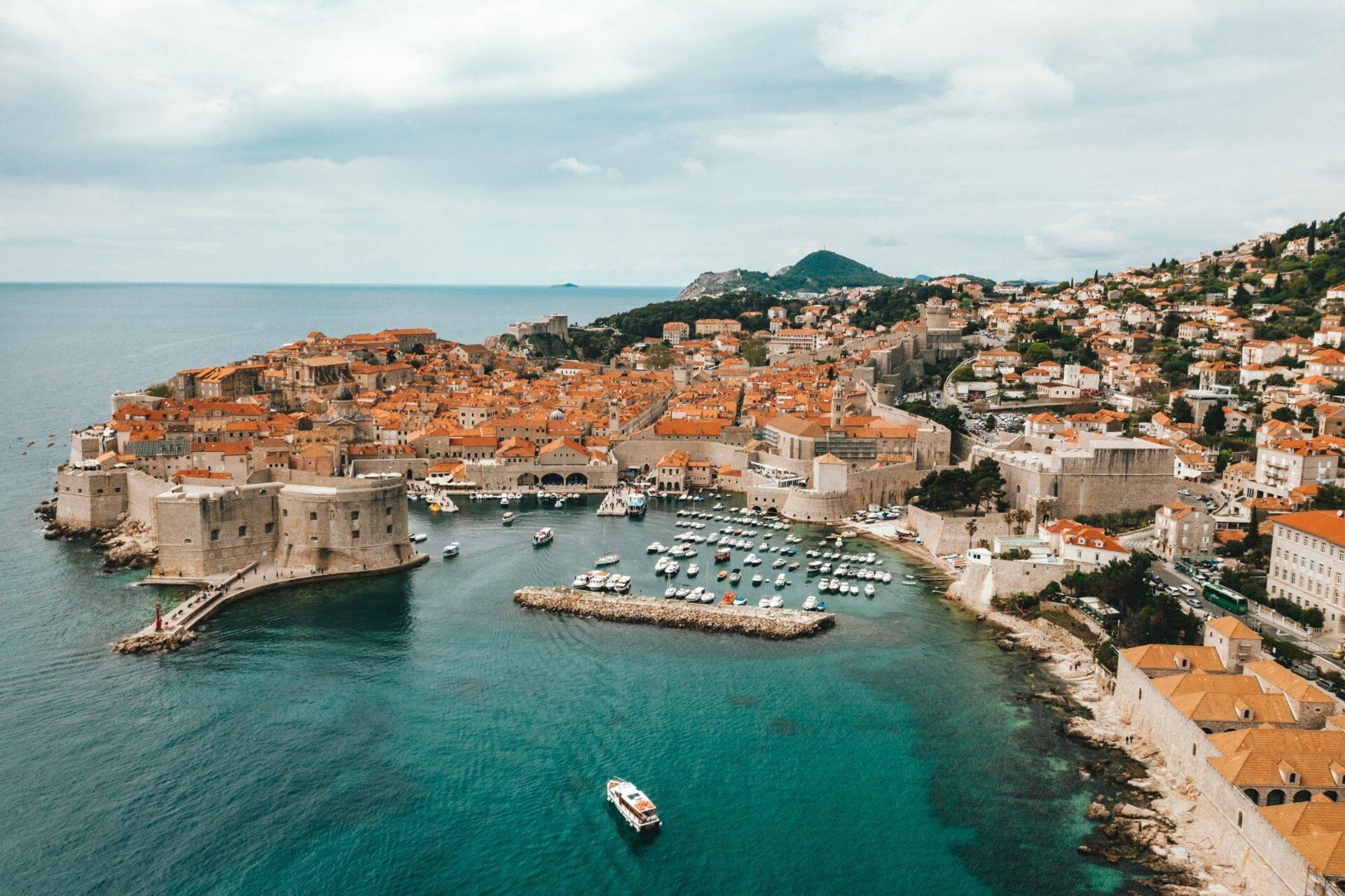 Vue aérienne d’une ville côtière fortifiée aux toits orangés avec port de plaisance et montagne en arrière-plan