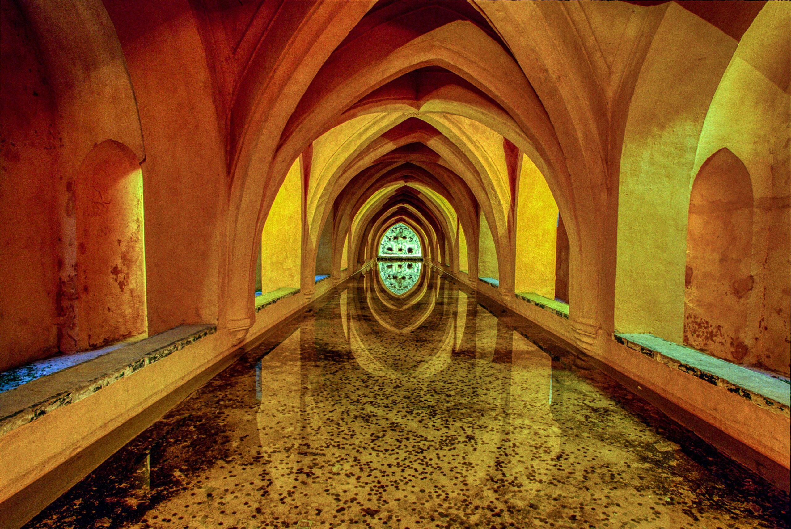 Galerie souterraine avec voûtes gothiques successives illuminées en jaune et rouge, sol inondé reflétant l’architecture
