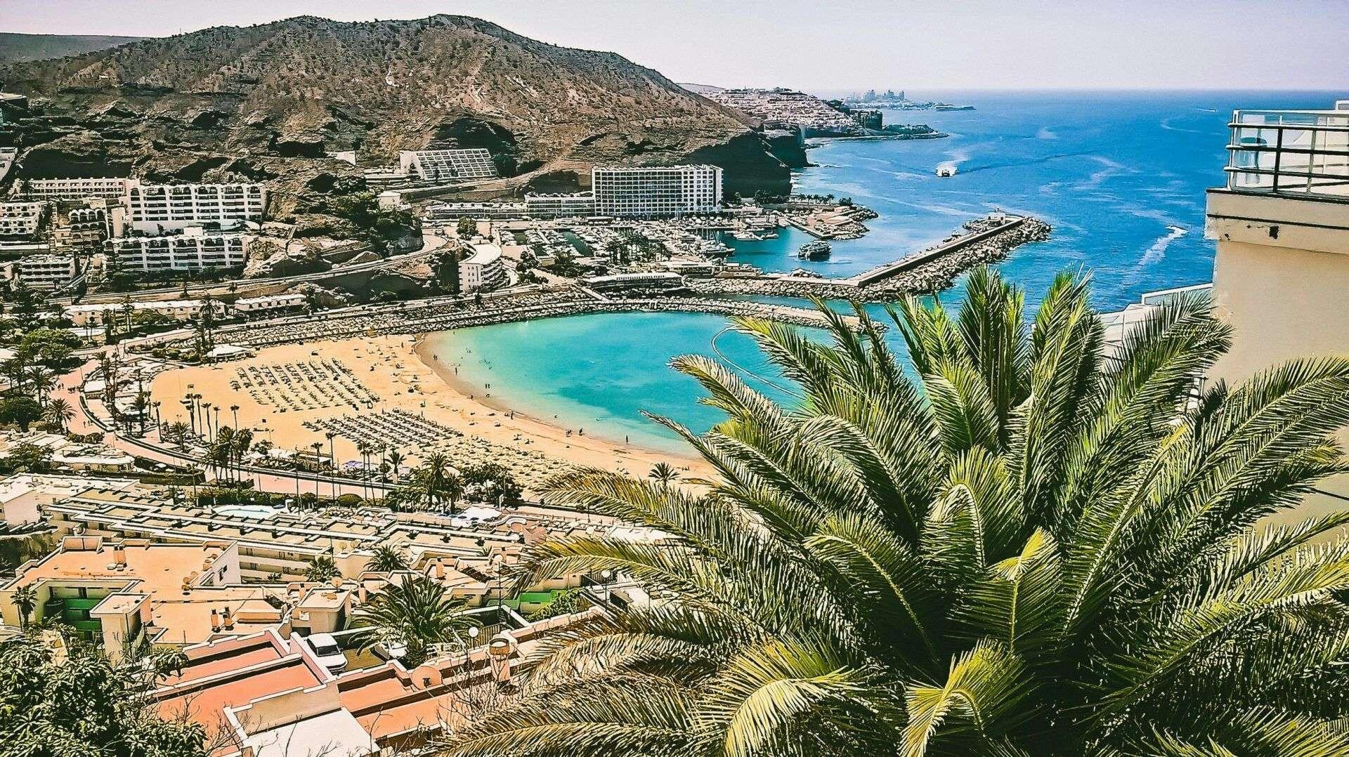 Vue panoramique d’une plage méditerranéenne avec hotels, montagnes rocheuses, eau turquoise et palmiers tropicaux