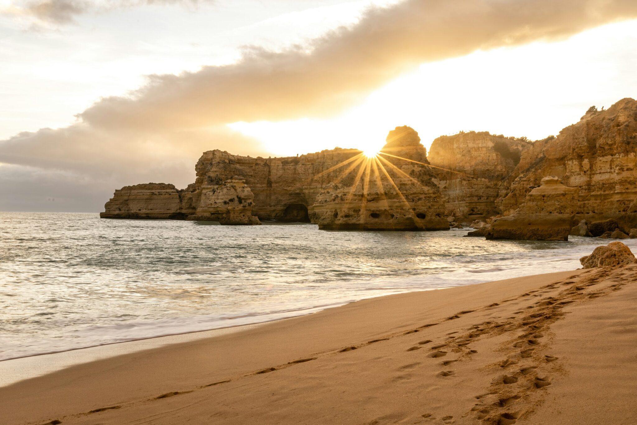 Plage dorée avec falaises rocheuses spectaculaires et soleil couchant illuminant l’océan et les formations géologiques