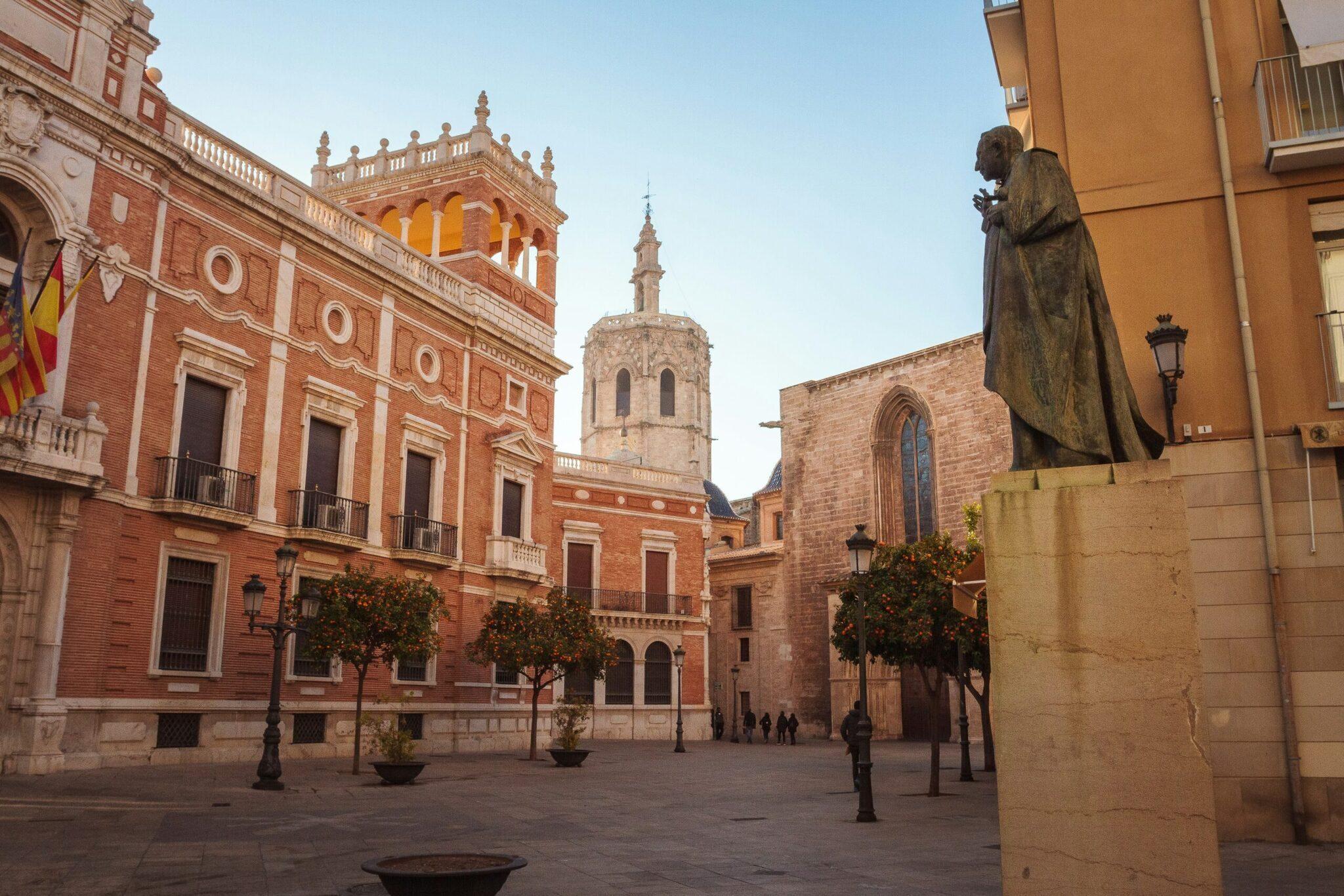 Une place historique en Espagne avec bâtiments en brique rouge, une statue et une église au fond, sous un ciel dégagé