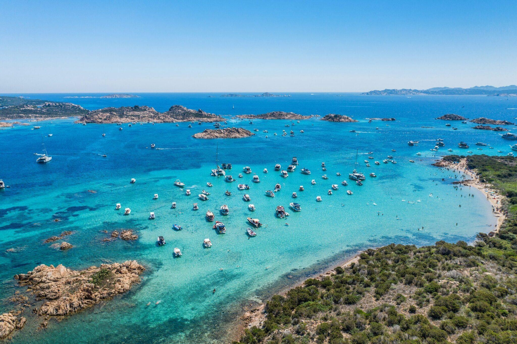 Aerial view of a turquoise bay dotted with moored boats, surrounded by rocky islets and lush Mediterranean coastline
