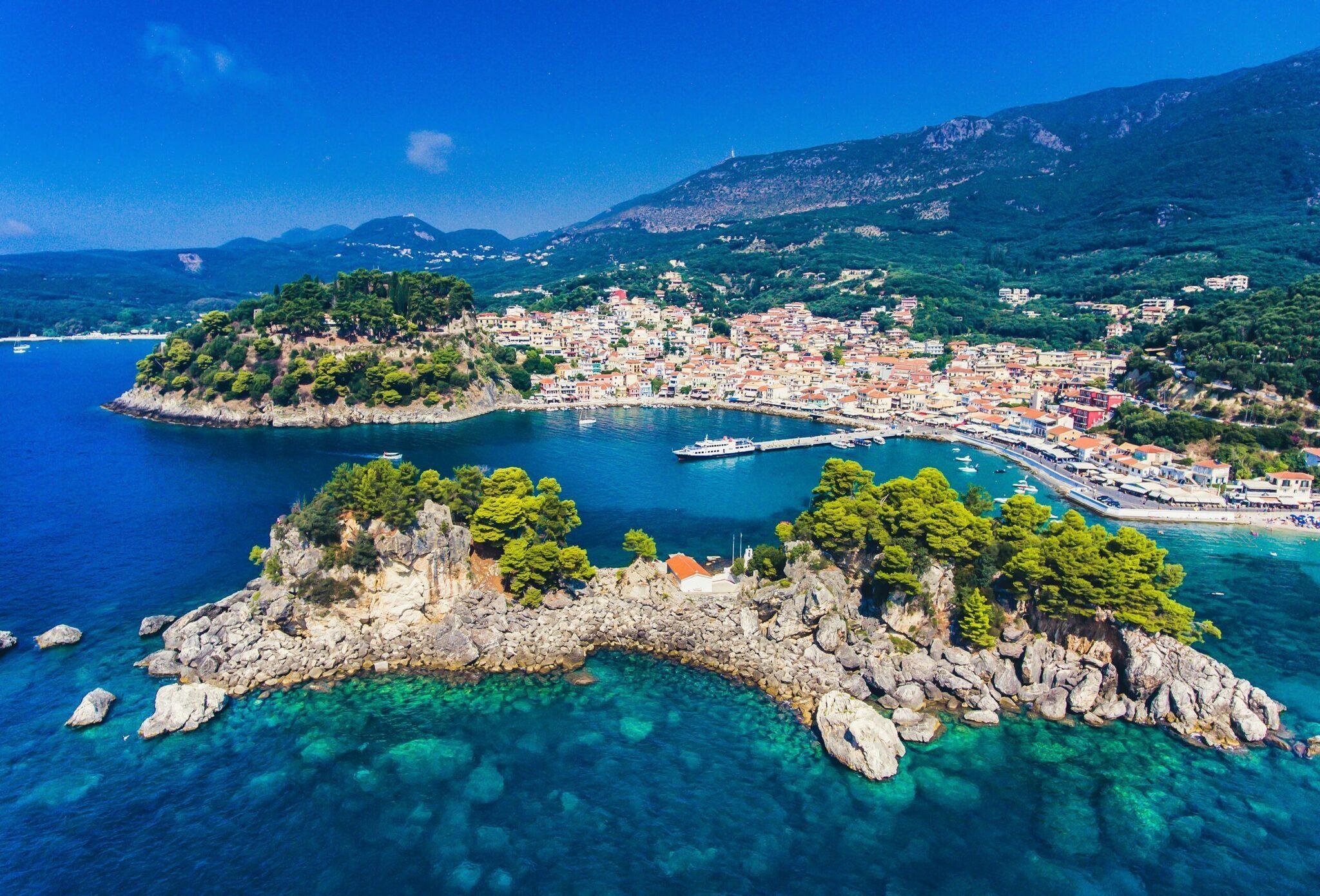 Aerial view of a Mediterranean bay with a rocky islet, a coastal town with orange rooftops, and crystal-clear turquoise waters