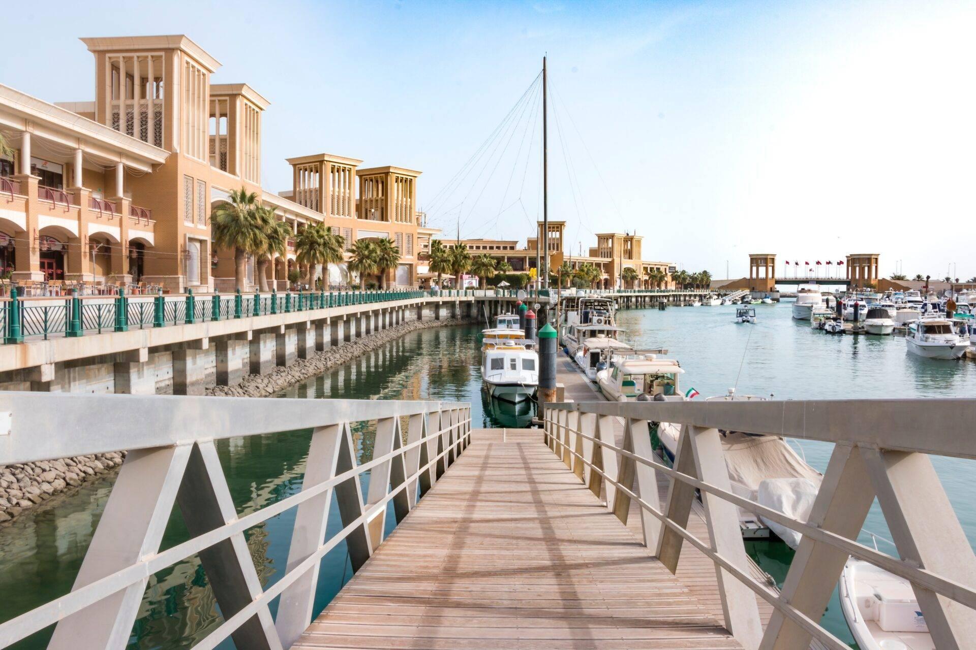 Un ponton en bois s’étend vers un port marina avec bateaux, bâtiments traditionnels beige et palmiers sous ciel dégagé