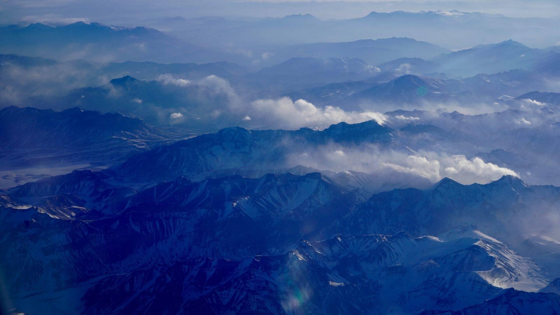 Vue aérienne d’une chaîne montagneuse majeure avec sommets enneigés, vallées profondes et nuages blancs dispersés