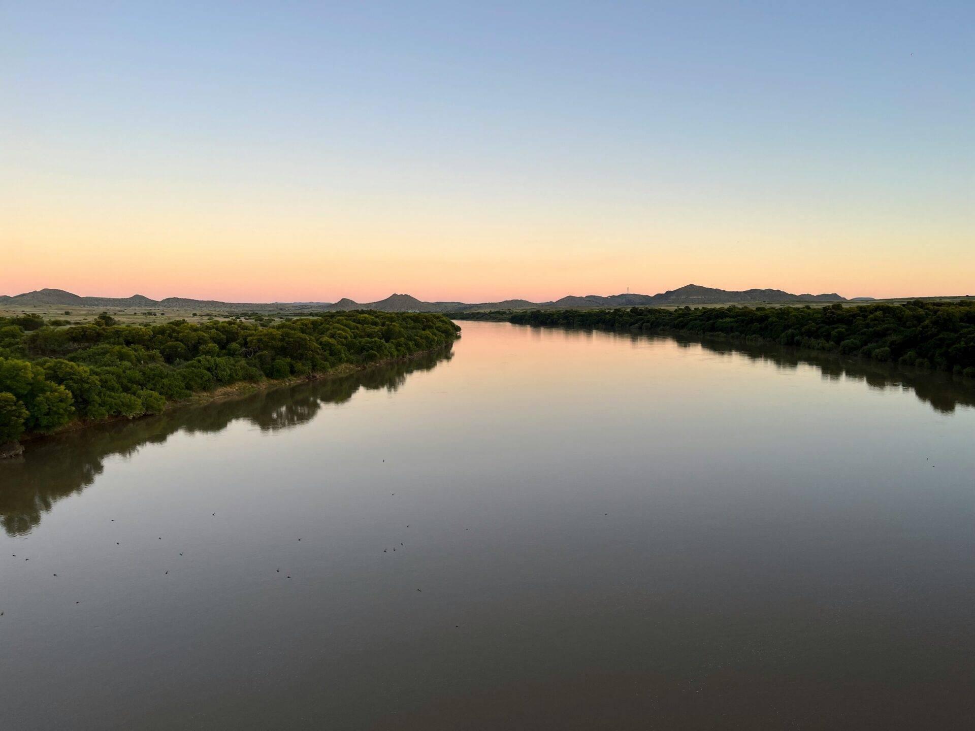 Un fleuve calme bordé de végétation luxuriante au crépuscule, avec des collines rocheuses en arrière-plan