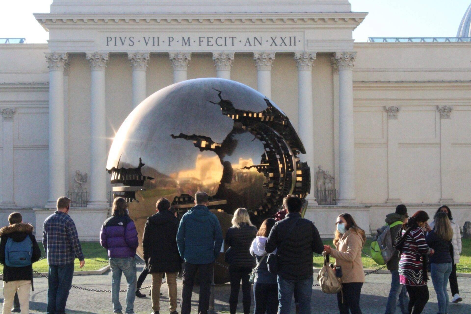 Un groupe de visiteurs observe une gigantesque sphère métallique réfléchissante devant un édifice classique aux colonnes blanches