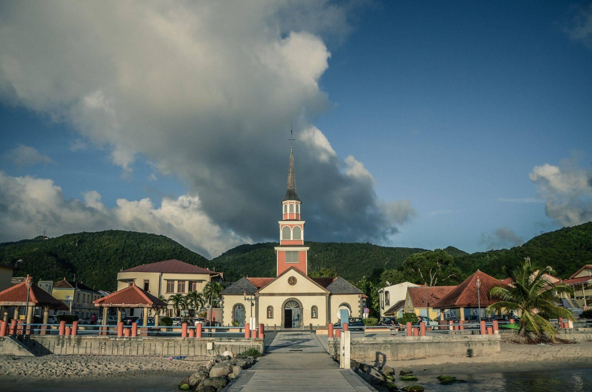 Église colorée en bord de mer avec clocher, entourée de maisons aux toits rouges et de montagnes verdoyantes