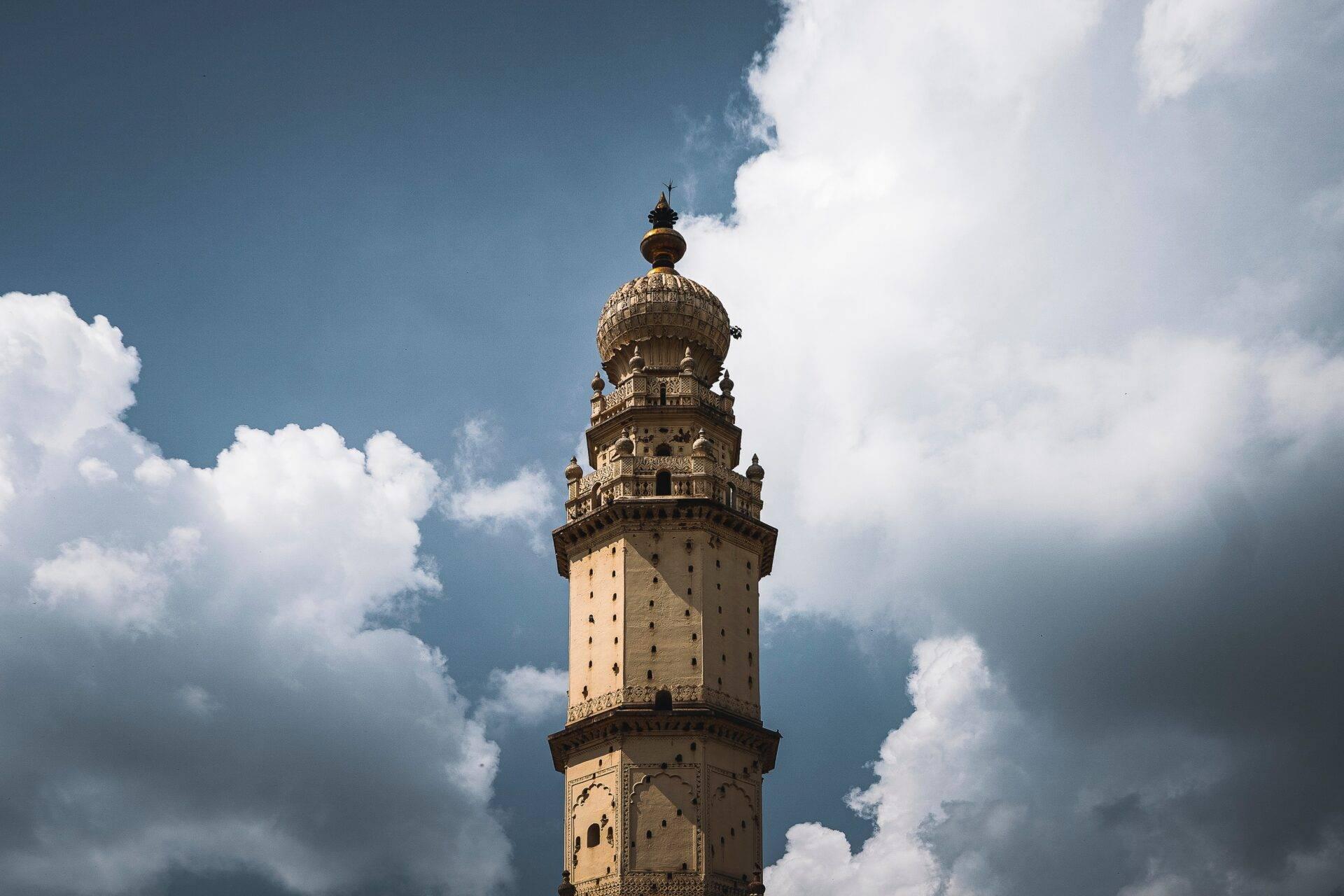 Minaret ancien en pierre ocre avec balcons ornementés sous un ciel bleu nuageux