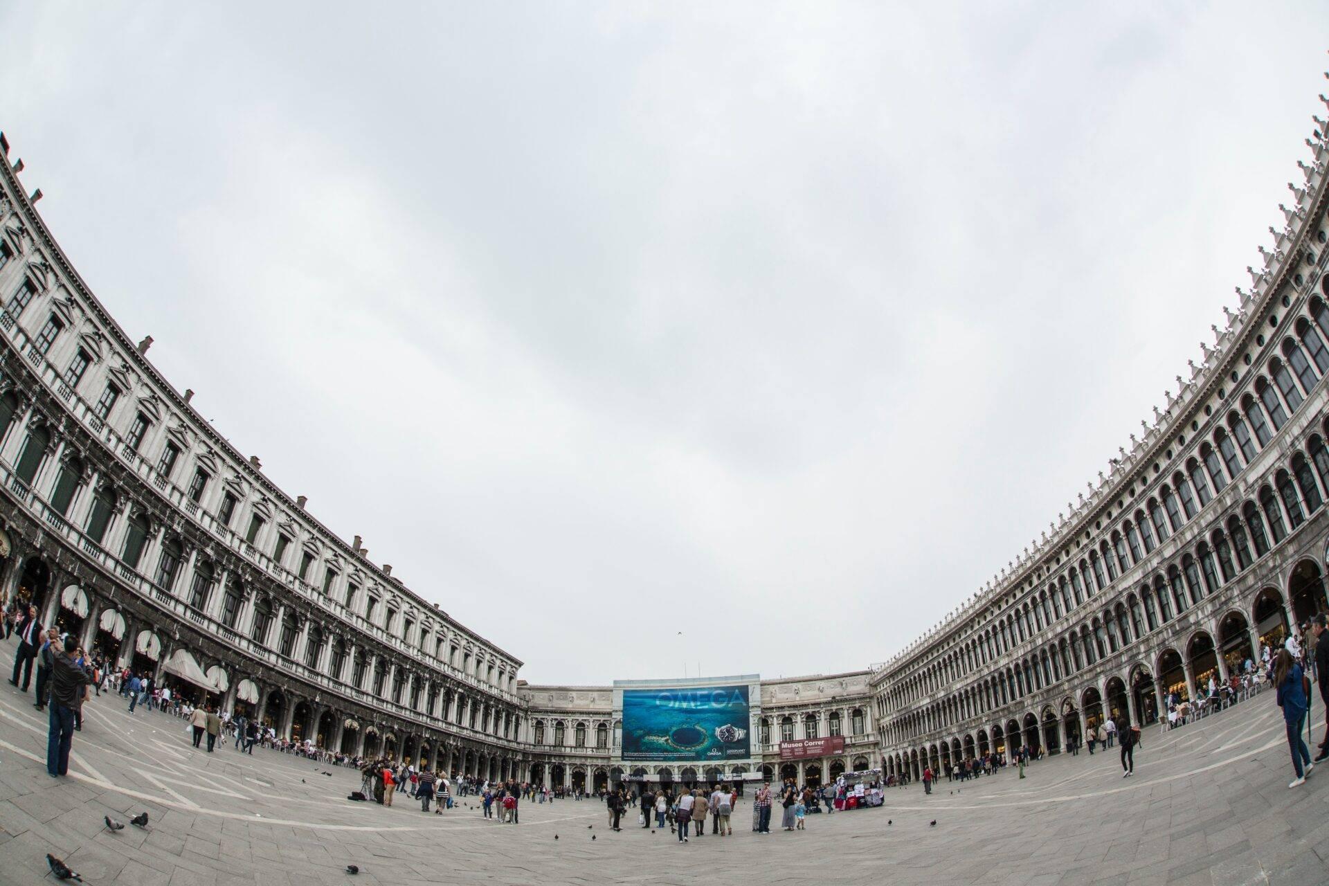 Une grande place carrée entourée d’élégants bâtiments historiques avec arcades, remplie de touristes