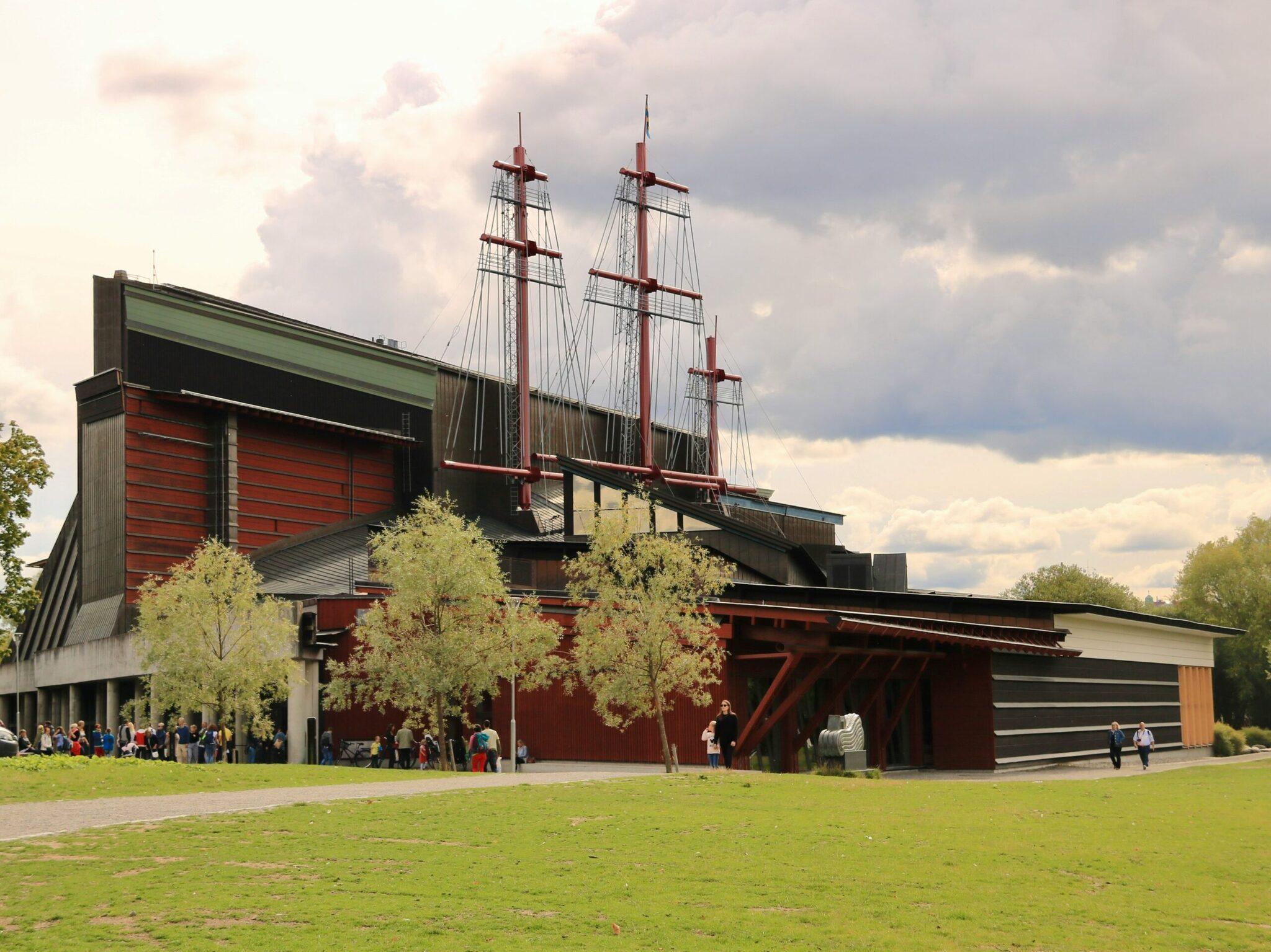 Un musée moderne aux façades rouges avec une réplique de navire à voiles intégré au toit, entouré de pelouses et d’arbres