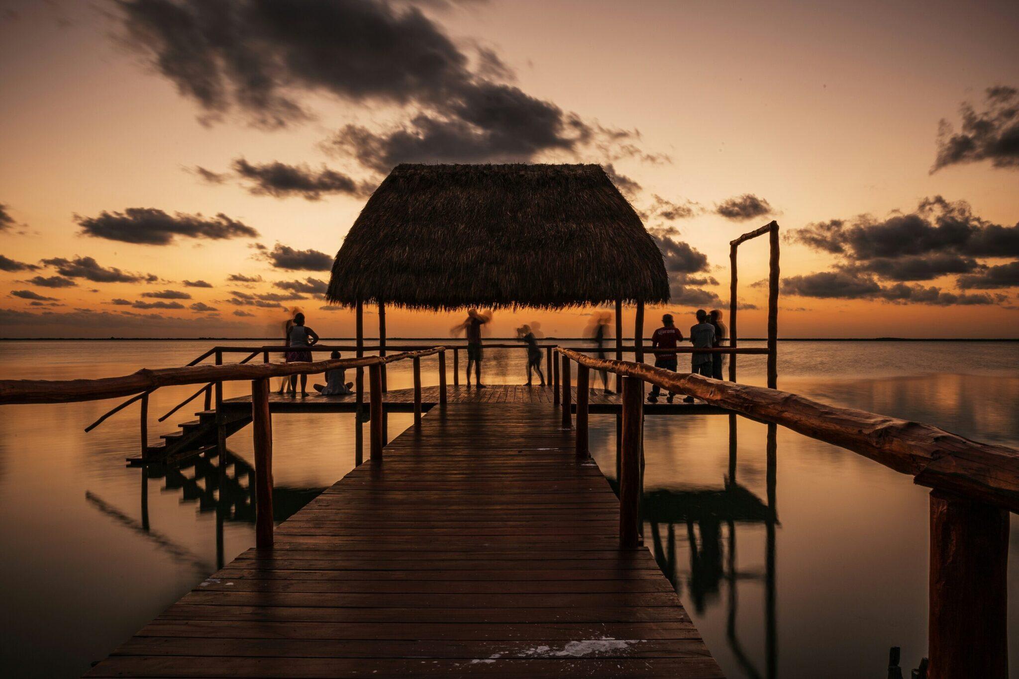 Un ponton en bois avec un abri à toit de chaume mène vers une baie calme au coucher du soleil