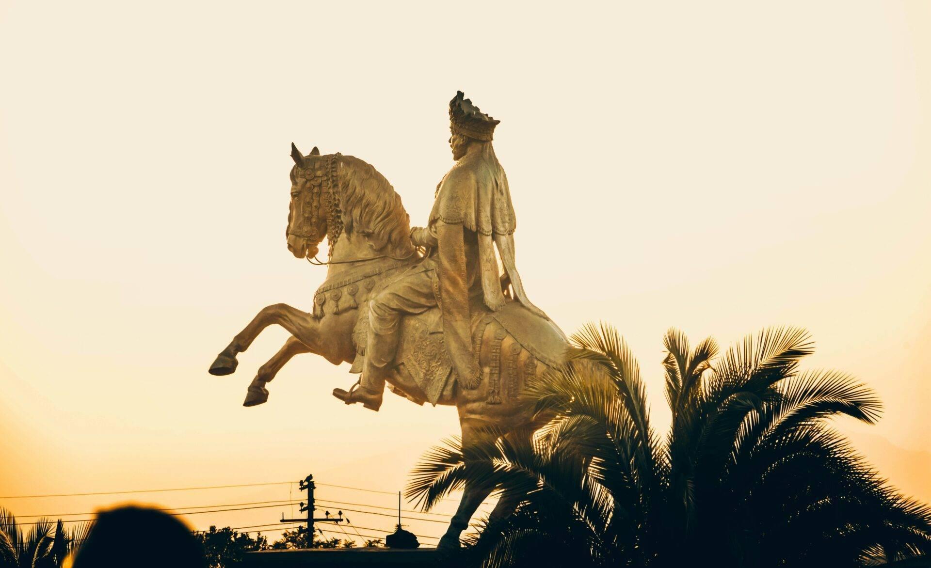 Statue dorée équestre d’un cavalier en tenue royale au coucher du soleil, entourée de palmiers