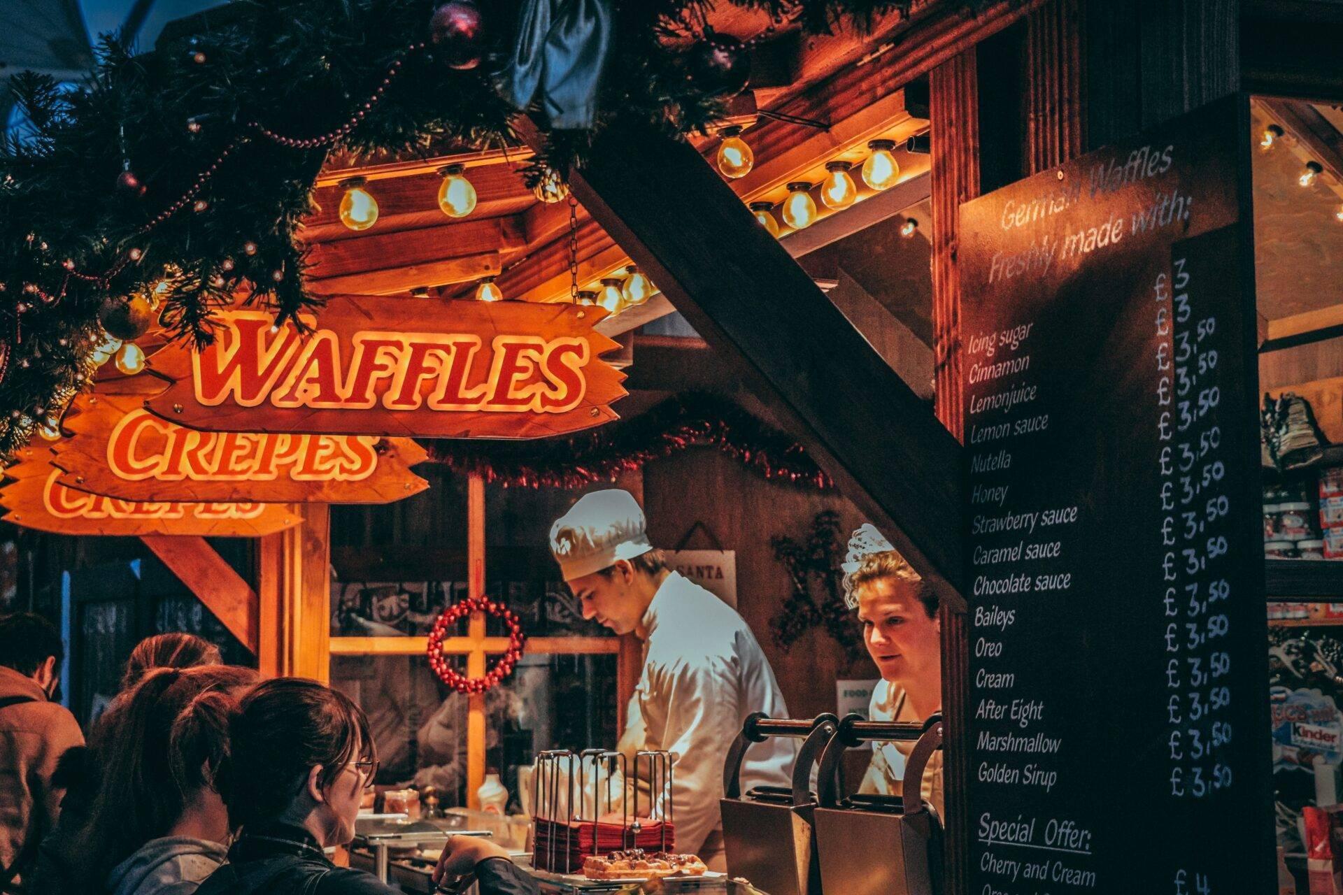 Un stand de crêpes et gaufres illuminé dans un marché de Noël européen avec menu affiché et vendeurs en action