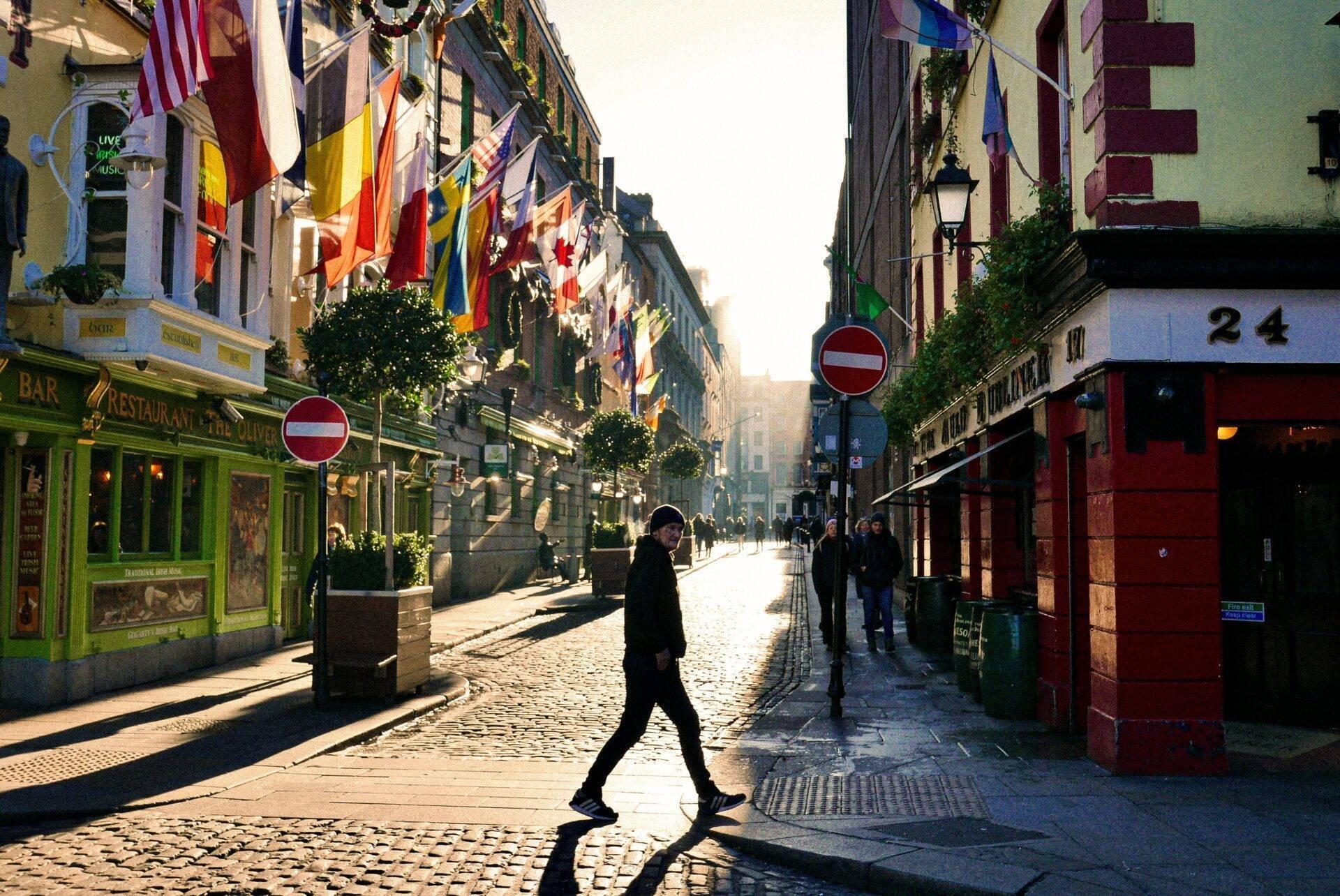 Une rue pavée européenne avec bars colorés, drapeaux internationaux et façades historiques, baignée de lumière naturelle