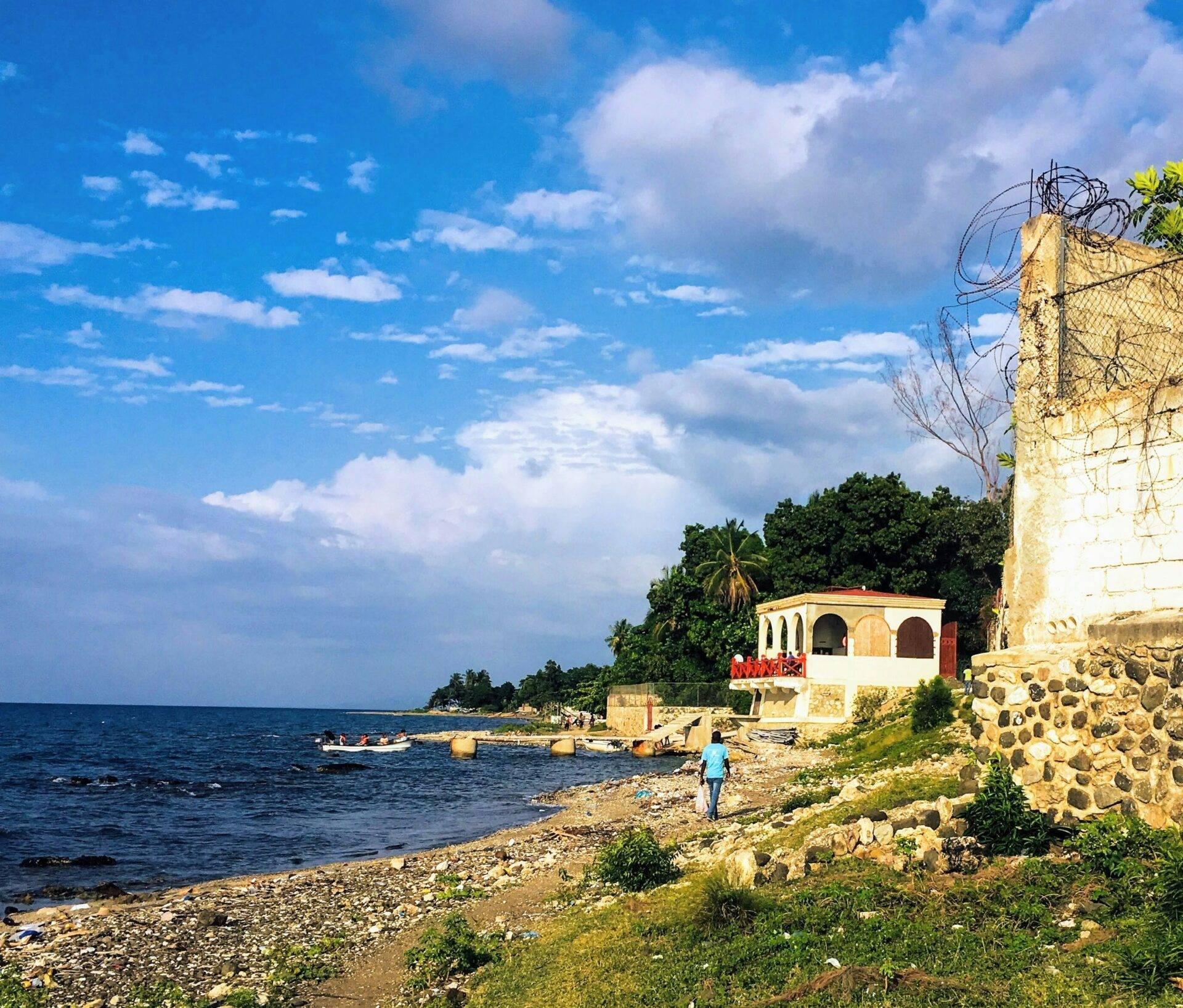 Une personne marche sur une plage rocheuse face à une maison colorée en pierre au bord de la mer avec des bateaux
