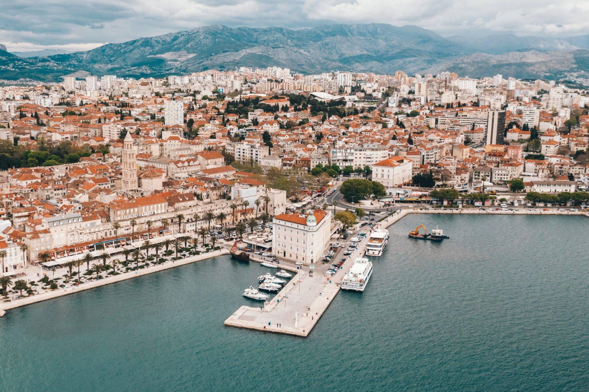 Vue aérienne d’une ville côtière méditerranéenne avec port, toits terracotta et montagnes verdoyantes en arrière-plan