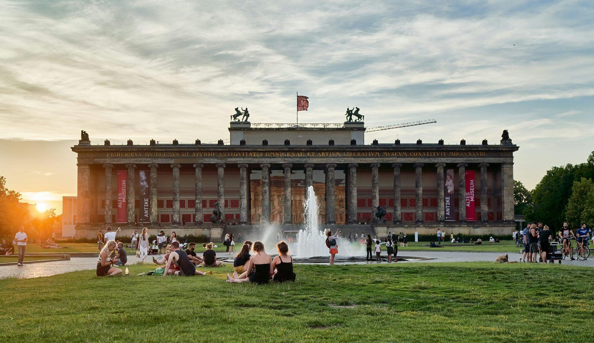 Un musée néoclassique avec colonnes imposantes et fontaine, entouré de visiteurs détendus sur la pelouse au coucher du soleil