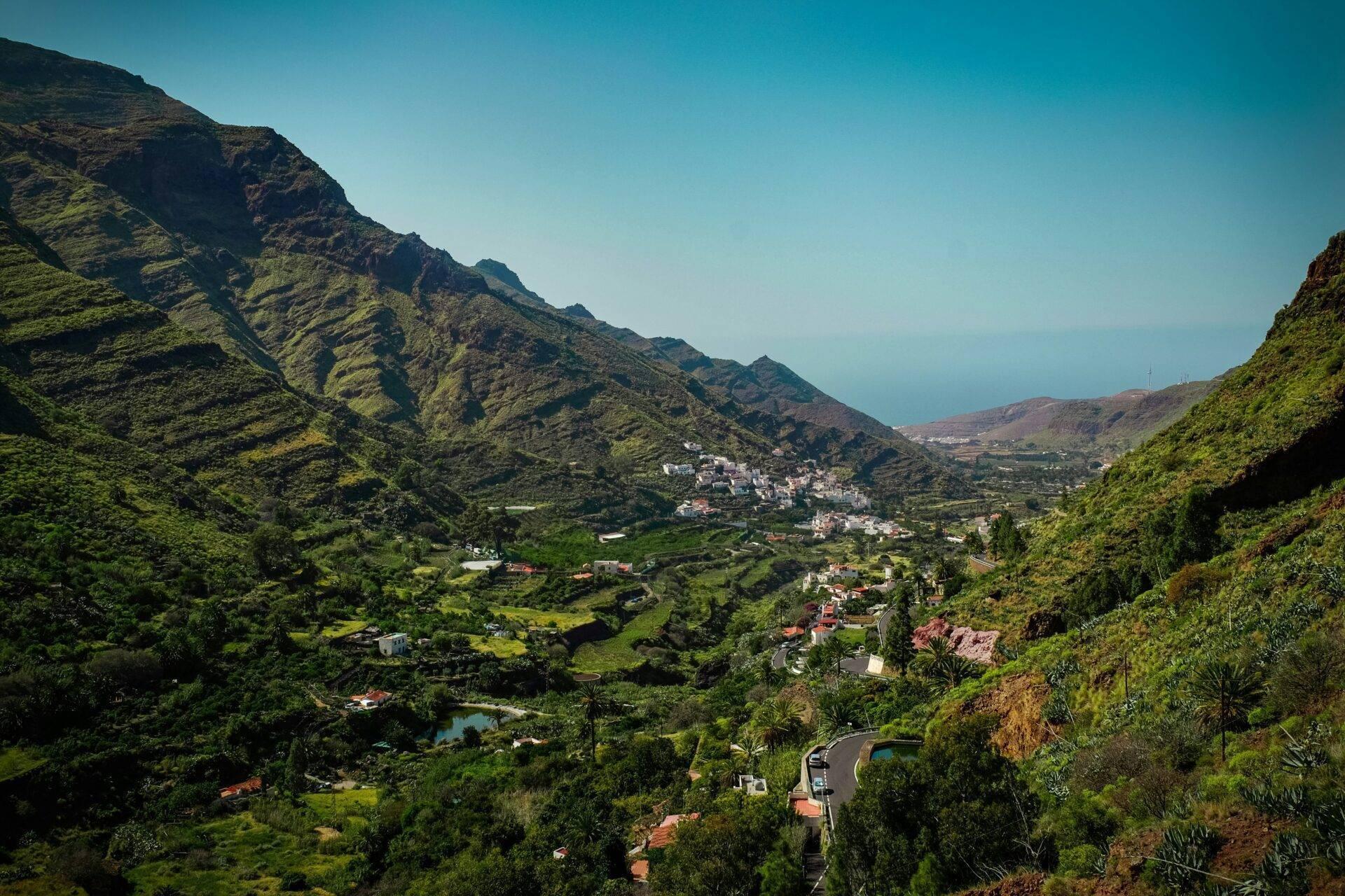 Vue aérienne d’une vallée verdoyante avec villages blancs, route sinueuse et montagnes escarpées sous ciel dégagé