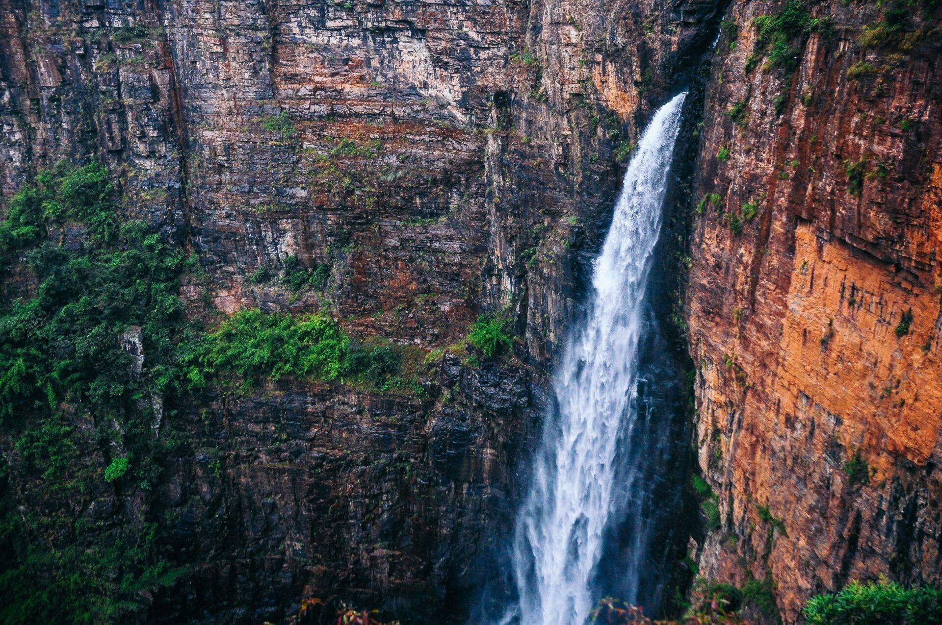Une cascade blanche dévale une falaise de roche rougeâtre encaissée, couverte de végétation verdoyante
