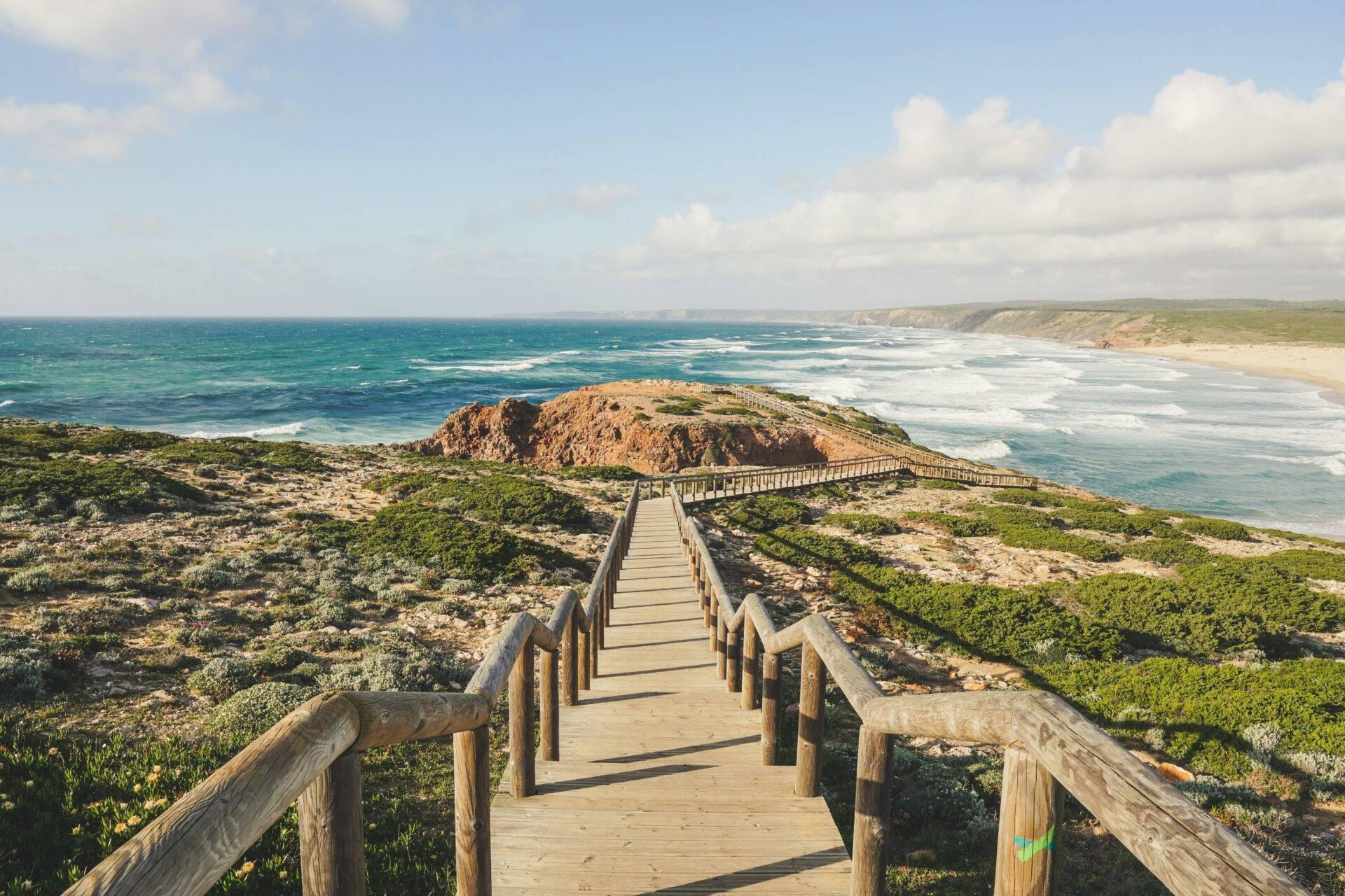 Passerelle en bois surplombant une côte rocheuse avec falaises verdoyantes et océan turquoise sous ciel dégagé
