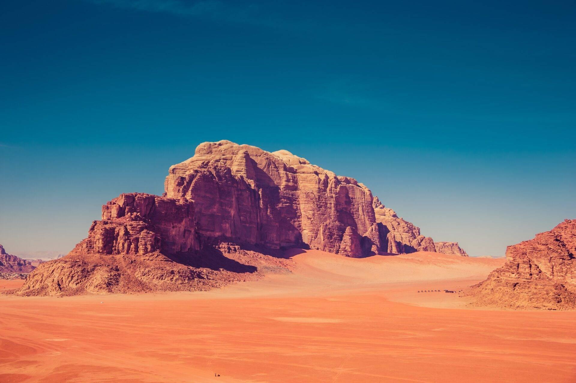 Un massif rocheux rouge ocre se dresse dans un désert de sable rougeâtre sous un ciel bleu dégagé
