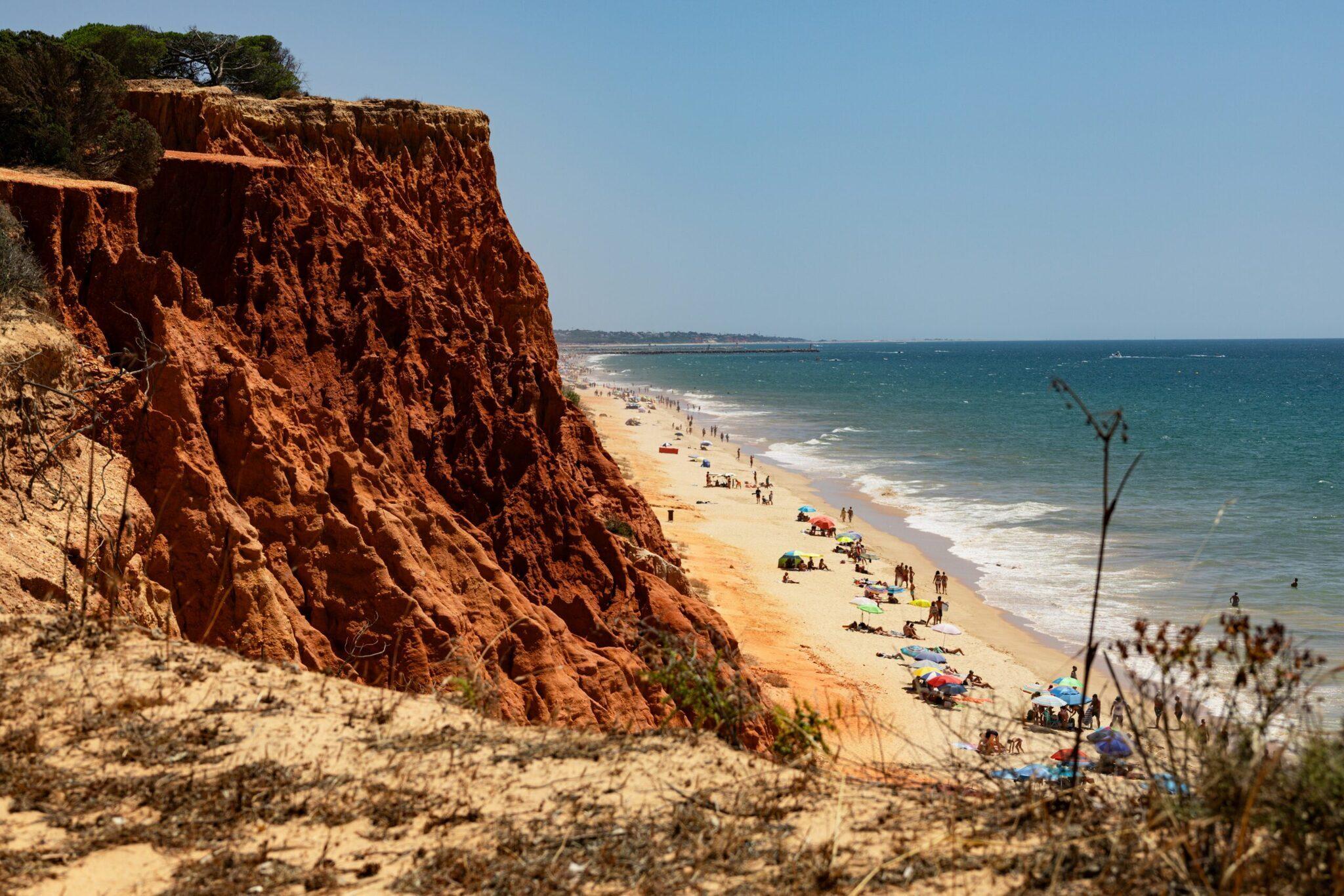 Falaise ocre surplombant une plage de sable doré avec baigneurs et parasols sous un ciel bleu dégagé