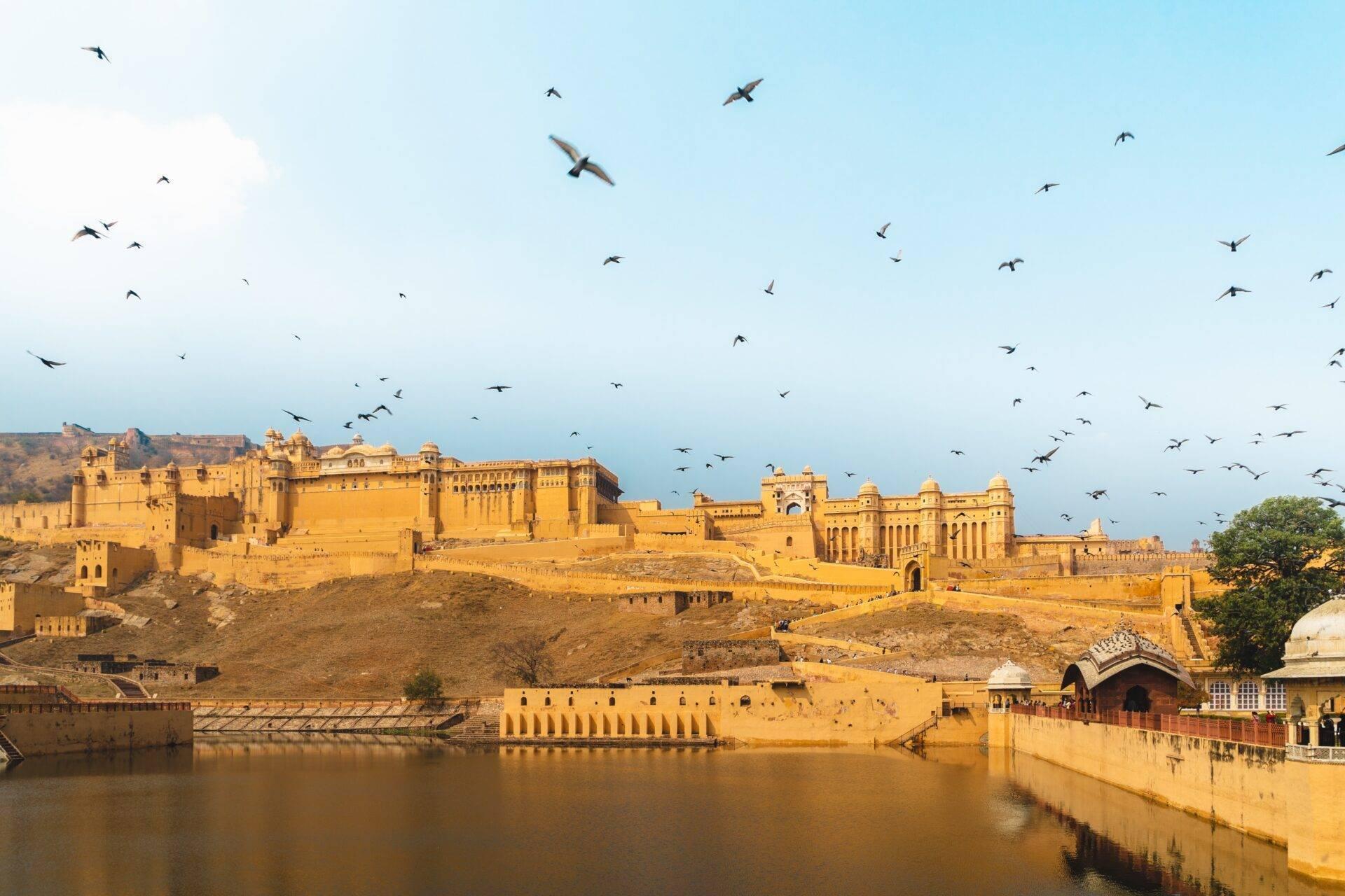 Un imposant fort en pierre ocre surplombant une rivière, avec des oiseaux en vol dans le ciel bleu