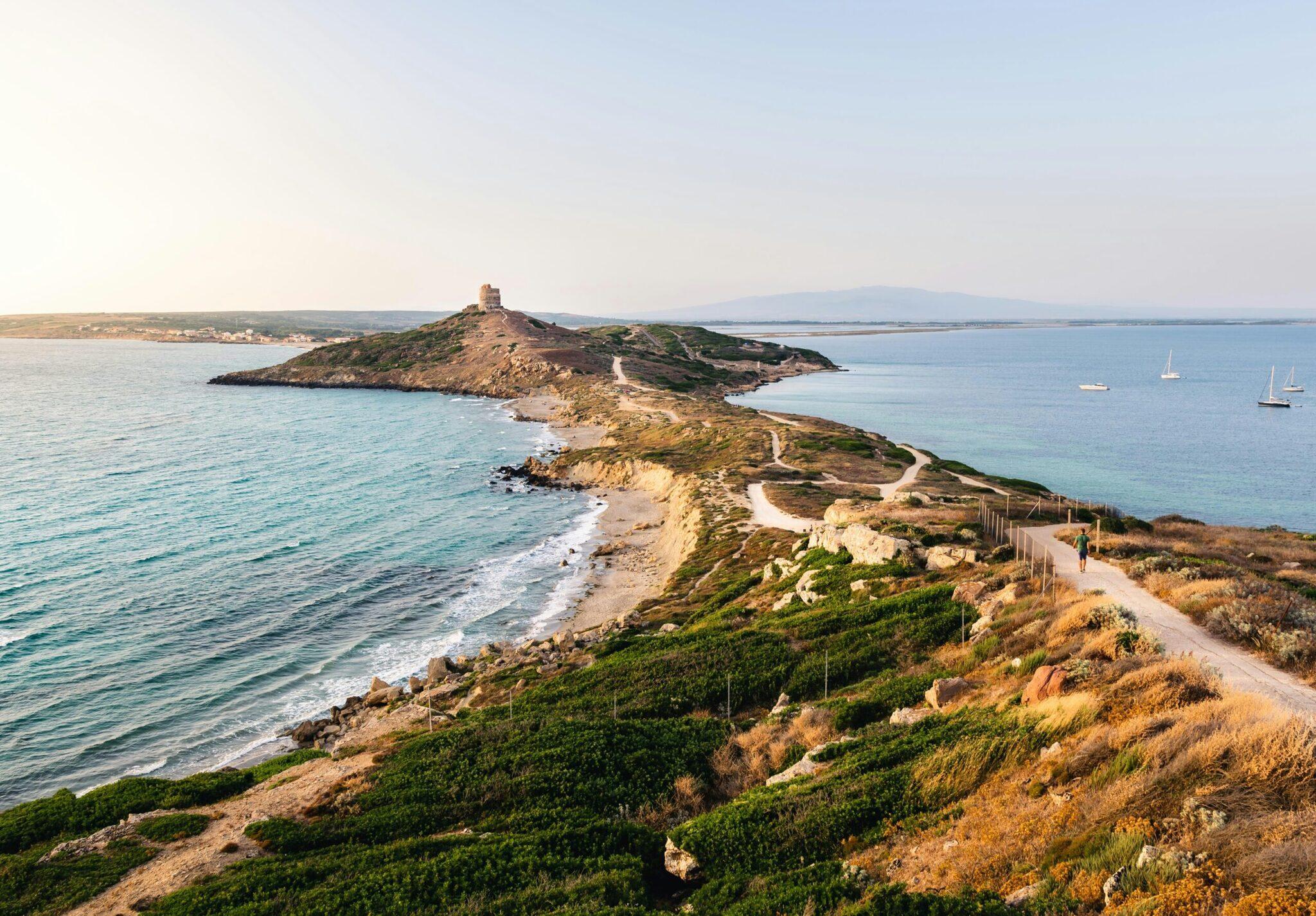 Winding coastal path above a turquoise bay with a fortified rocky islet and sailing boats at anchor