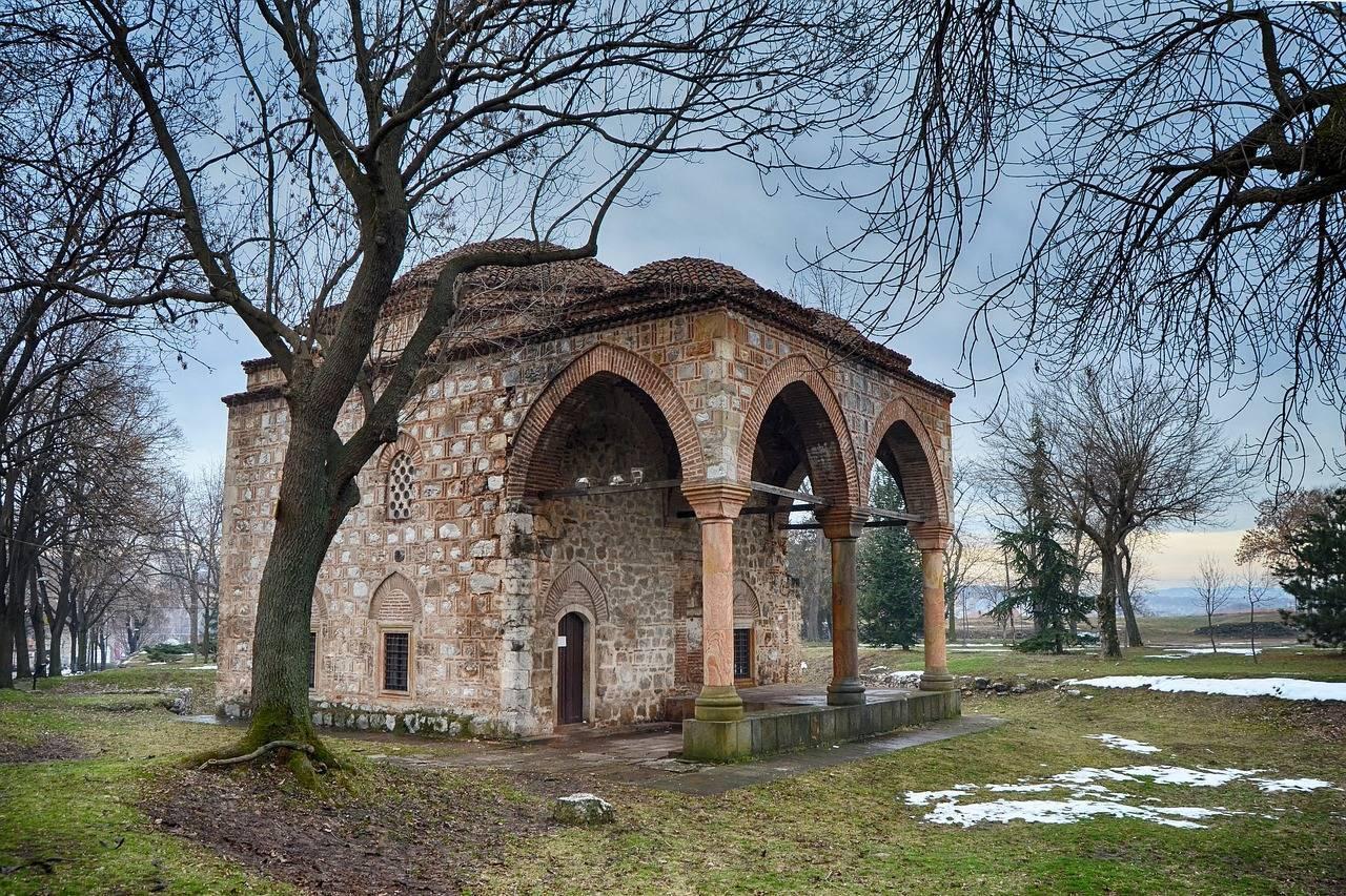 Un édifice religieux historique en pierre rouge avec arcades, entouré d’arbres nus et de prairies
